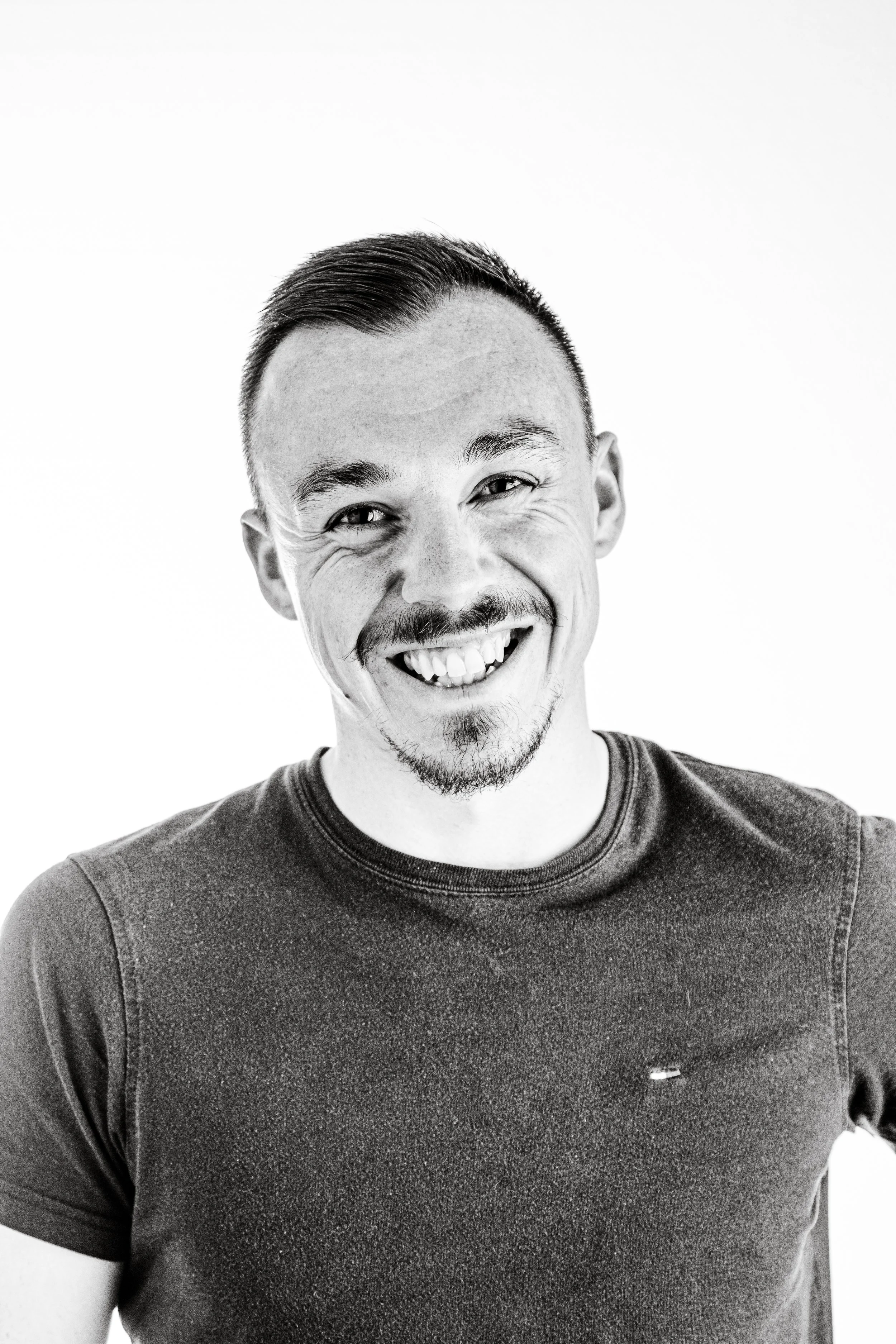 Black and white photo of a man with short hair, a mustache, and goatee, smiling with teeth showing, wearing a dark T-shirt, against a plain light background.