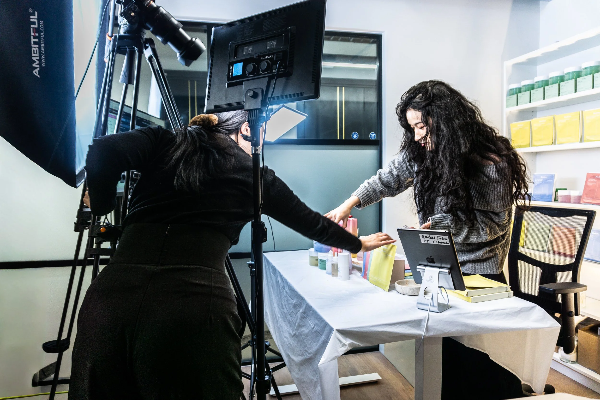 Two women setting up products on a table for a photoshoot in a well-lit room with shelves of colorful boxes in the background.
