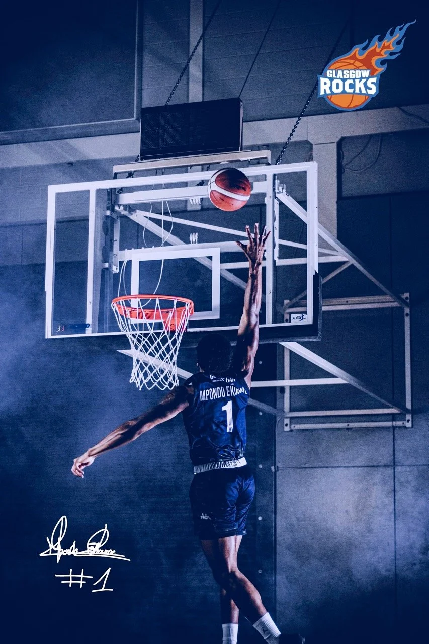 A basketball player in a dark blue uniform jumps to make a shot on an indoor basketball court. The player is reaching towards the basketball hoop with the ball just above the rim. There is a Glasgow Rocks logo in the top right corner.