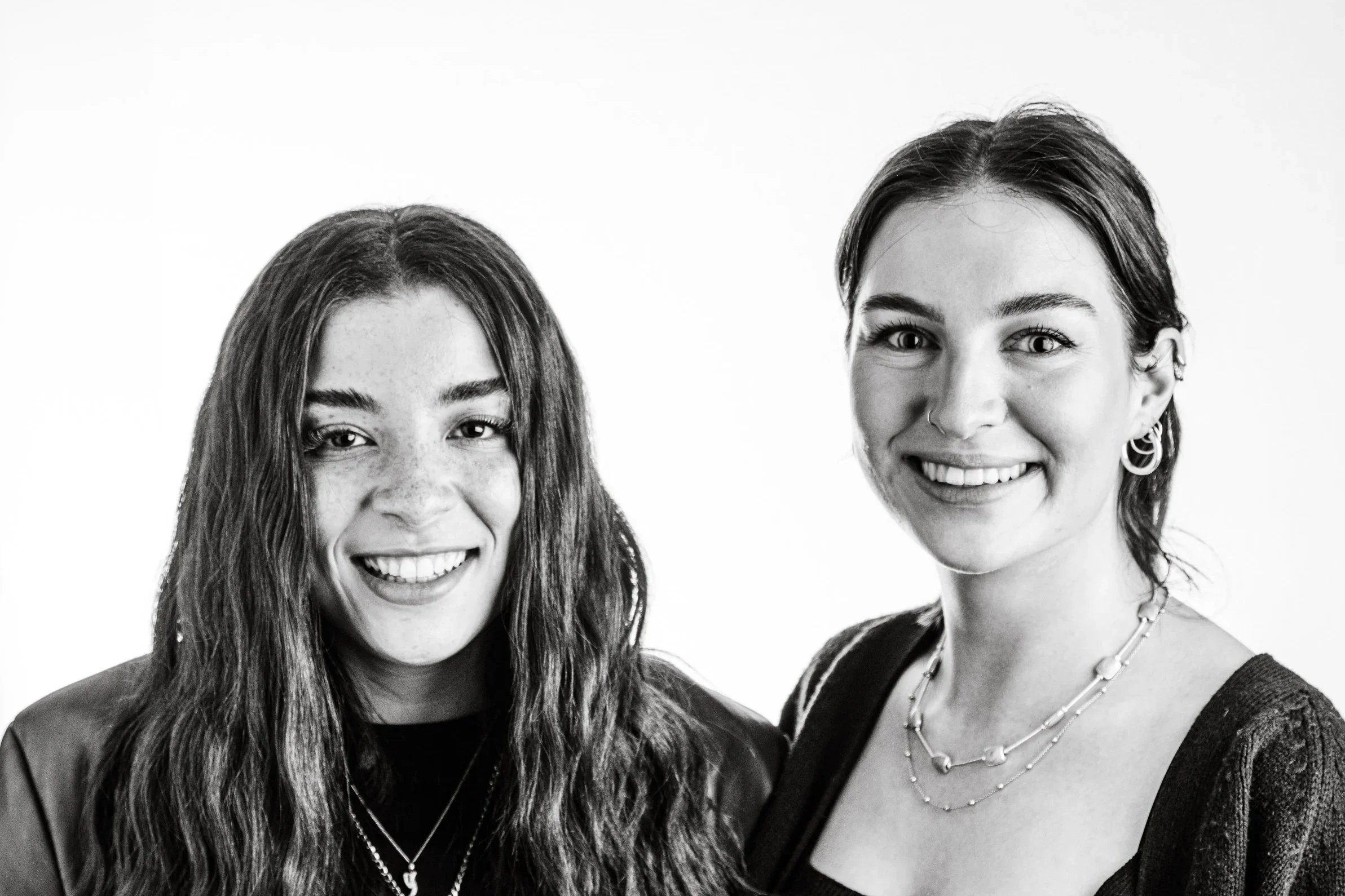 Black and white photo of two smiling women with long hair, one with glasses, earrings, necklaces, and casual clothing.