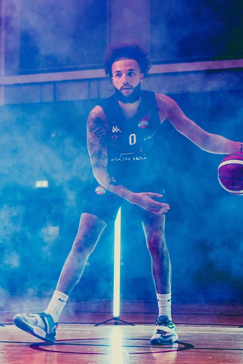 A basketball player with a beard and tattoos dribbling a ball on a gym court with fog and mist surrounding him, glowing blue light in the background.