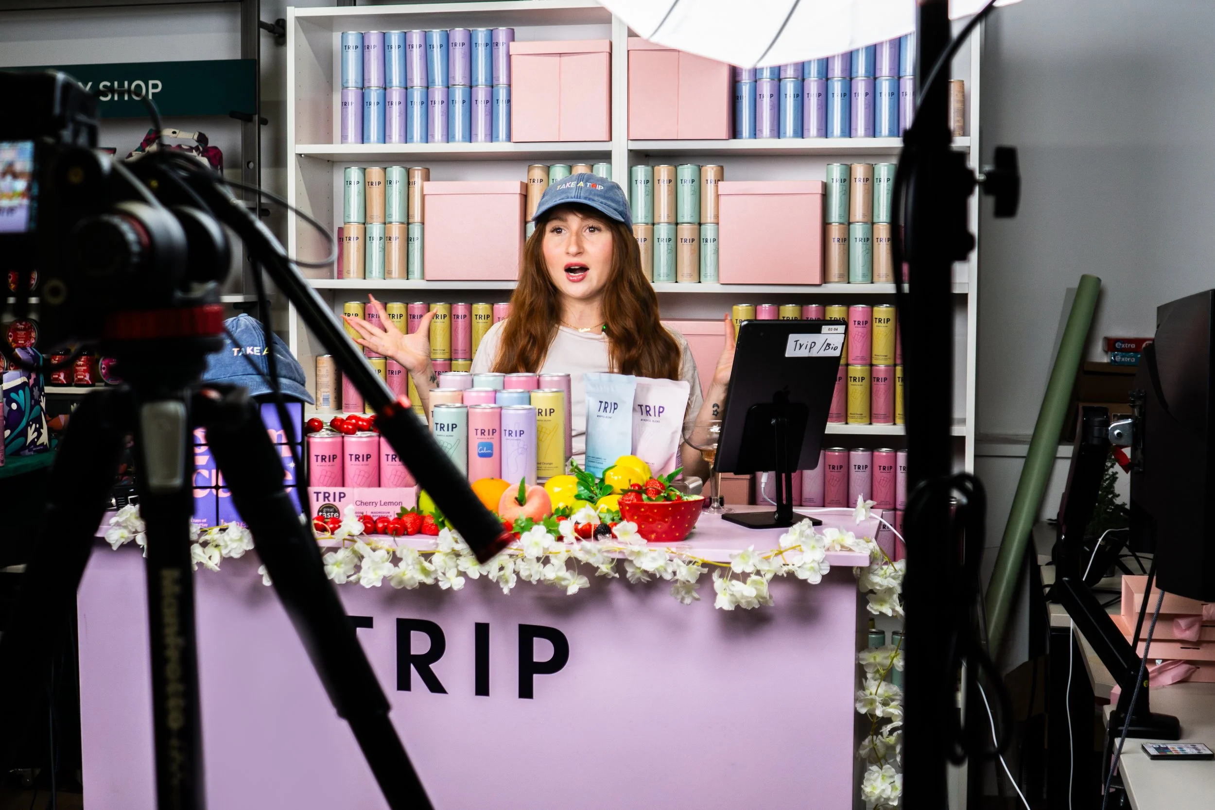 A woman at a pink booth decorated with flowers, surrounded by products labeled 'TRIP' and various fruits, filming a video.