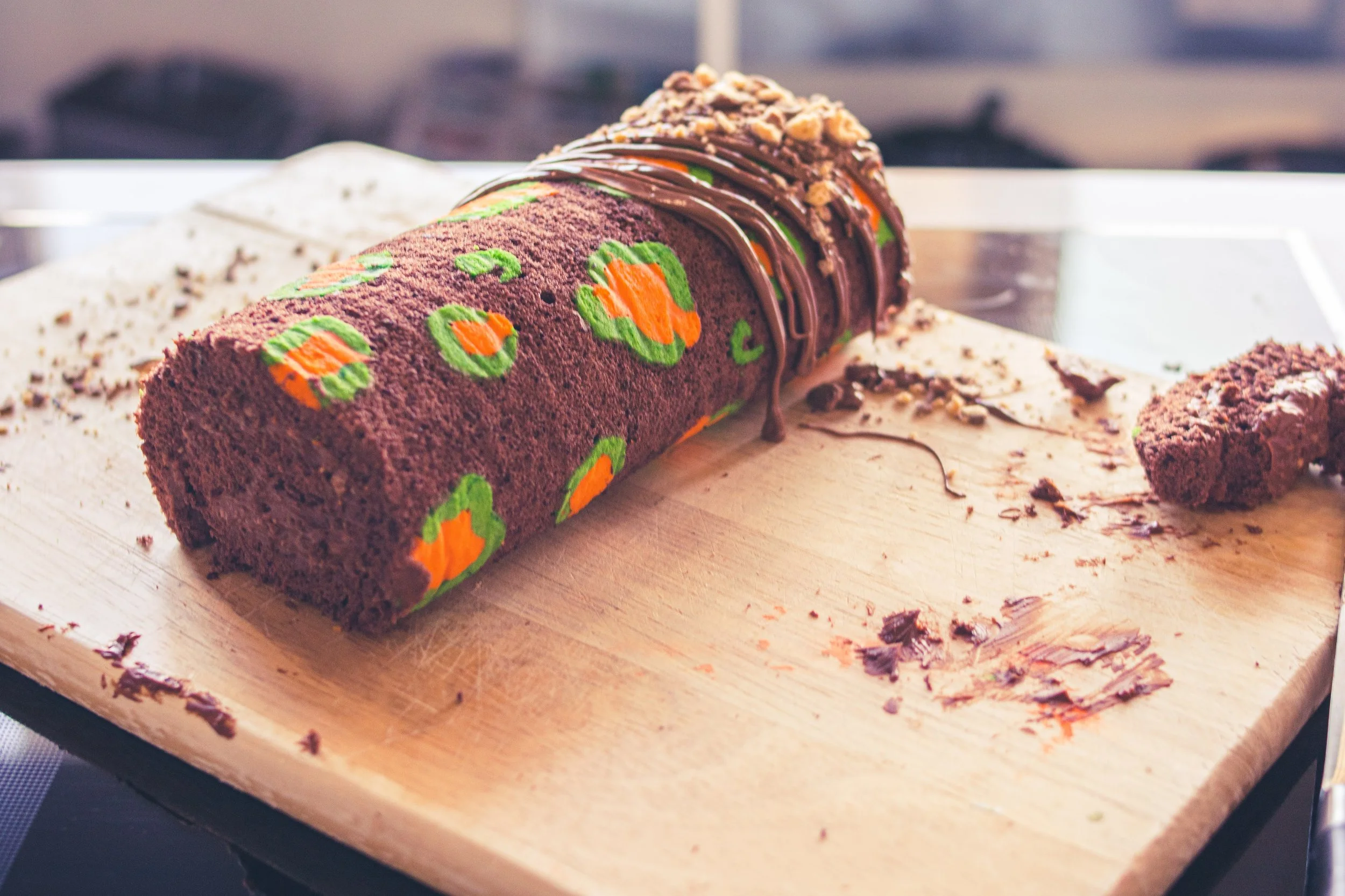 Chocolate Swiss roll cake with colorful pumpkin patterns, decorated with chocolate drizzle and chopped nuts on top, on a wooden cutting board with remnants of chocolate.