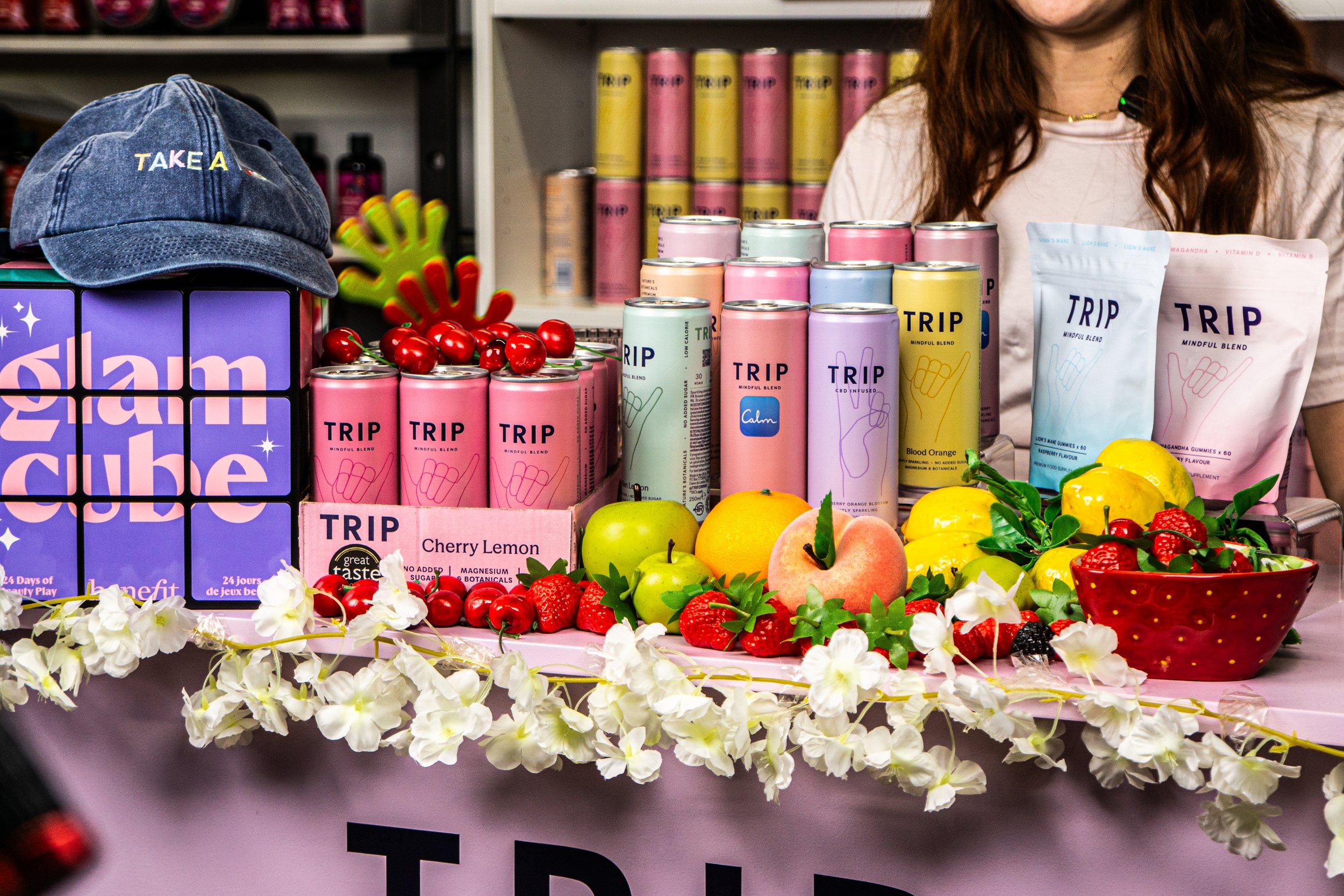 Display of colorful canned beverages labeled 'TRIP' with fresh fruits including cherries, strawberries, apples, peaches, and lemons arranged on a table decorated with white flowers. A woman is partially visible behind the display.