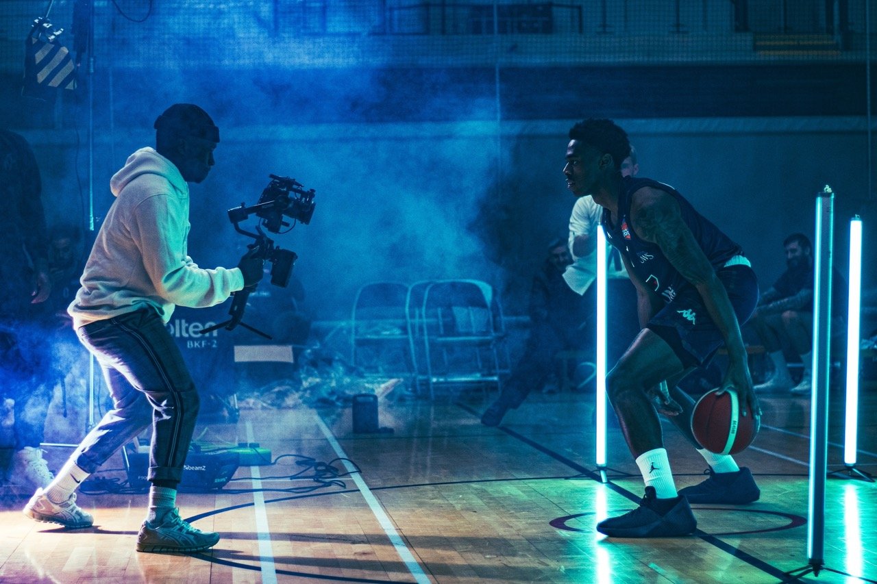 A basketball player holding a ball in a gym with blue lighting, while a person films with a camera. There are spectators in the background.