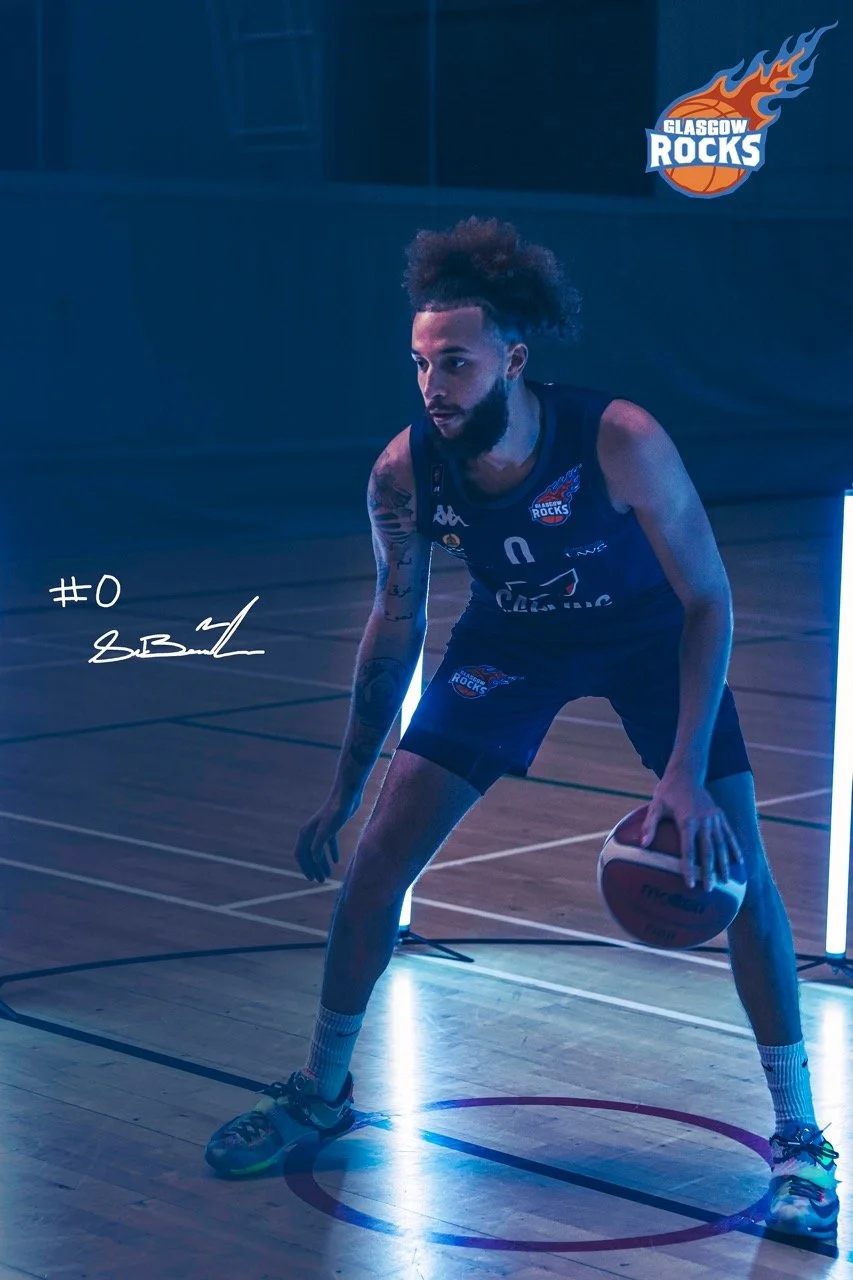 A basketball player in a dark uniform with the Glasgow Rocks logo is holding a ball on a basketball court. The scene is illuminated with blue lighting and features the Glasgow Rocks logo in the top right corner.