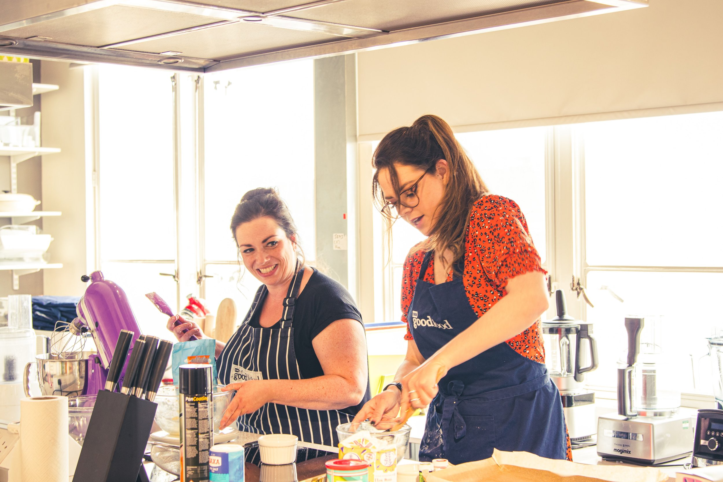 Two women baking in a bright kitchen, one smiling and looking at the camera, the other focused on mixing ingredients, with baking supplies and kitchen appliances on the counter.