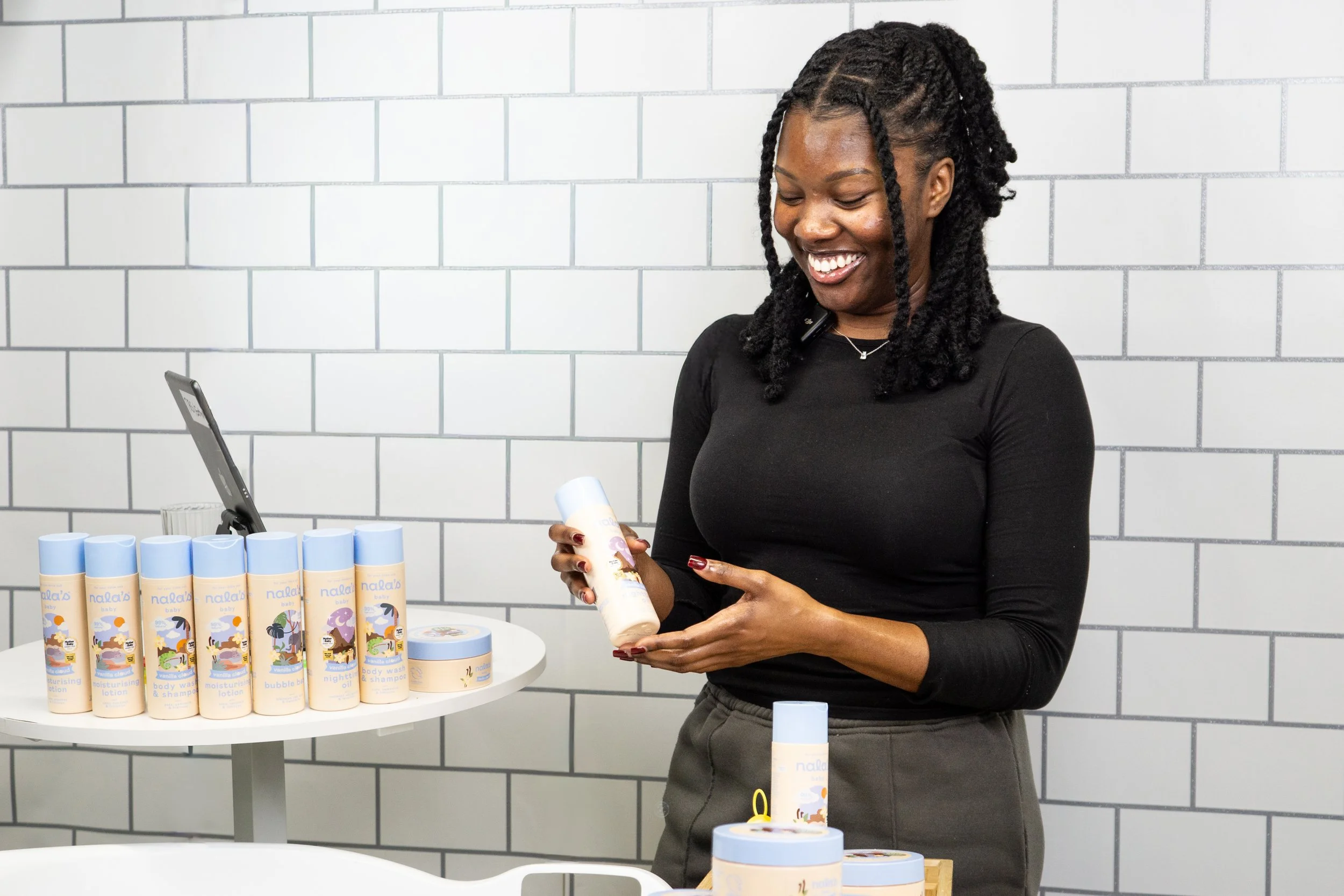A smiling woman in a black long-sleeve shirt displays a bottle of skincare or body care product in front of a display of similar bottles on a white round table against a white tiled wall.