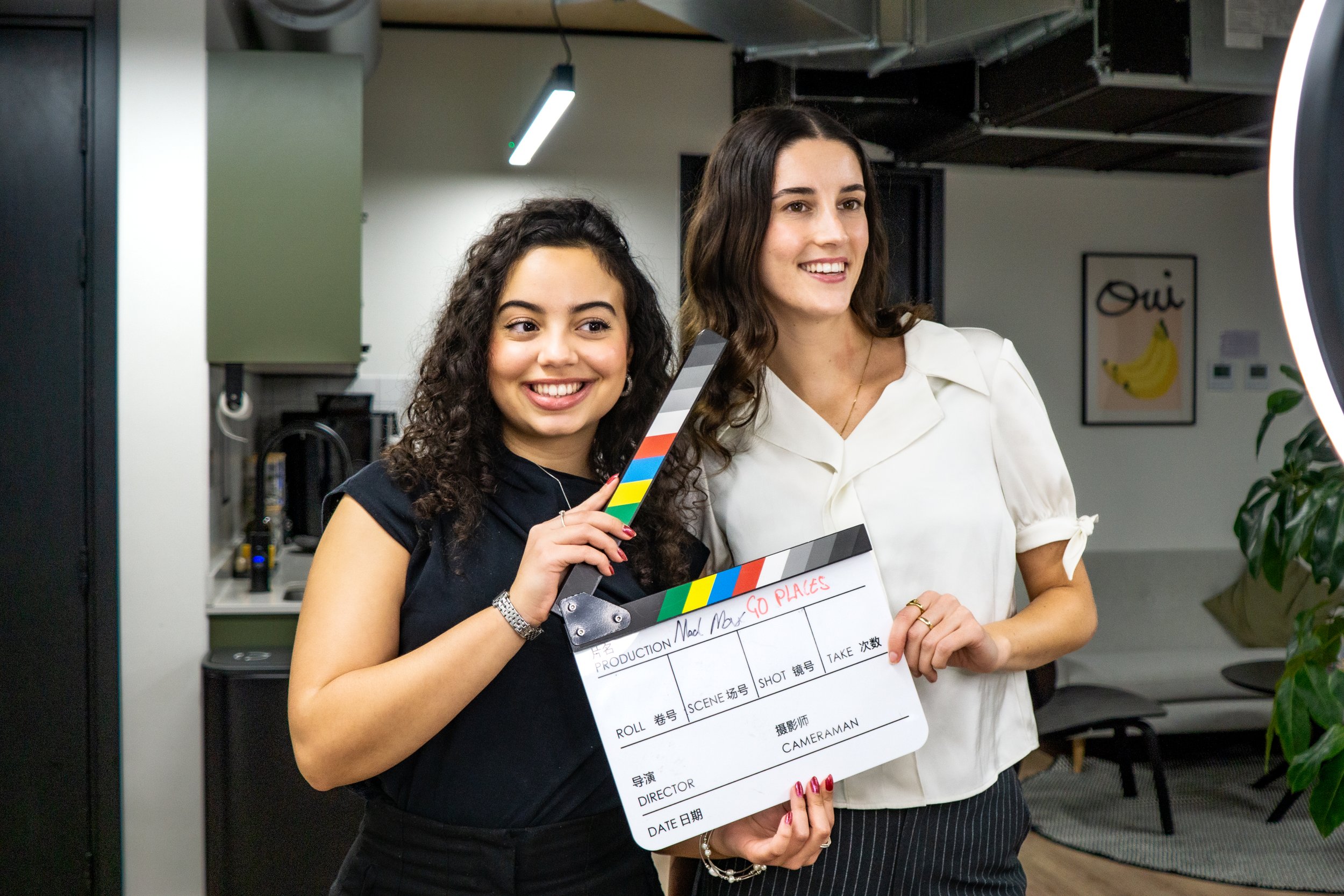 Two women on a film set, one holding a clapperboard and a film slate in a modern interior with studio lighting.
