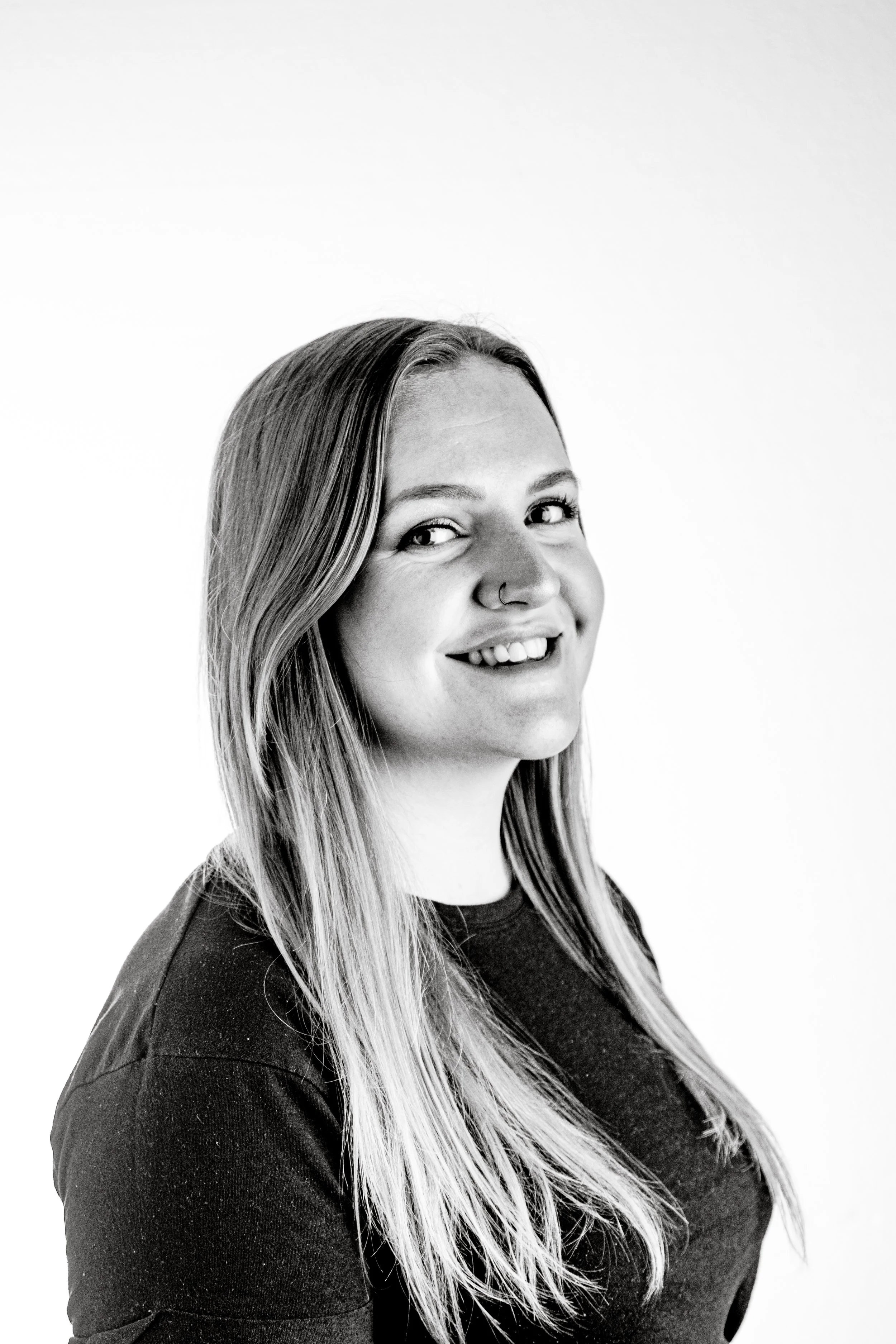 A smiling woman with long hair and a nose ring, looking at the camera, in black and white.