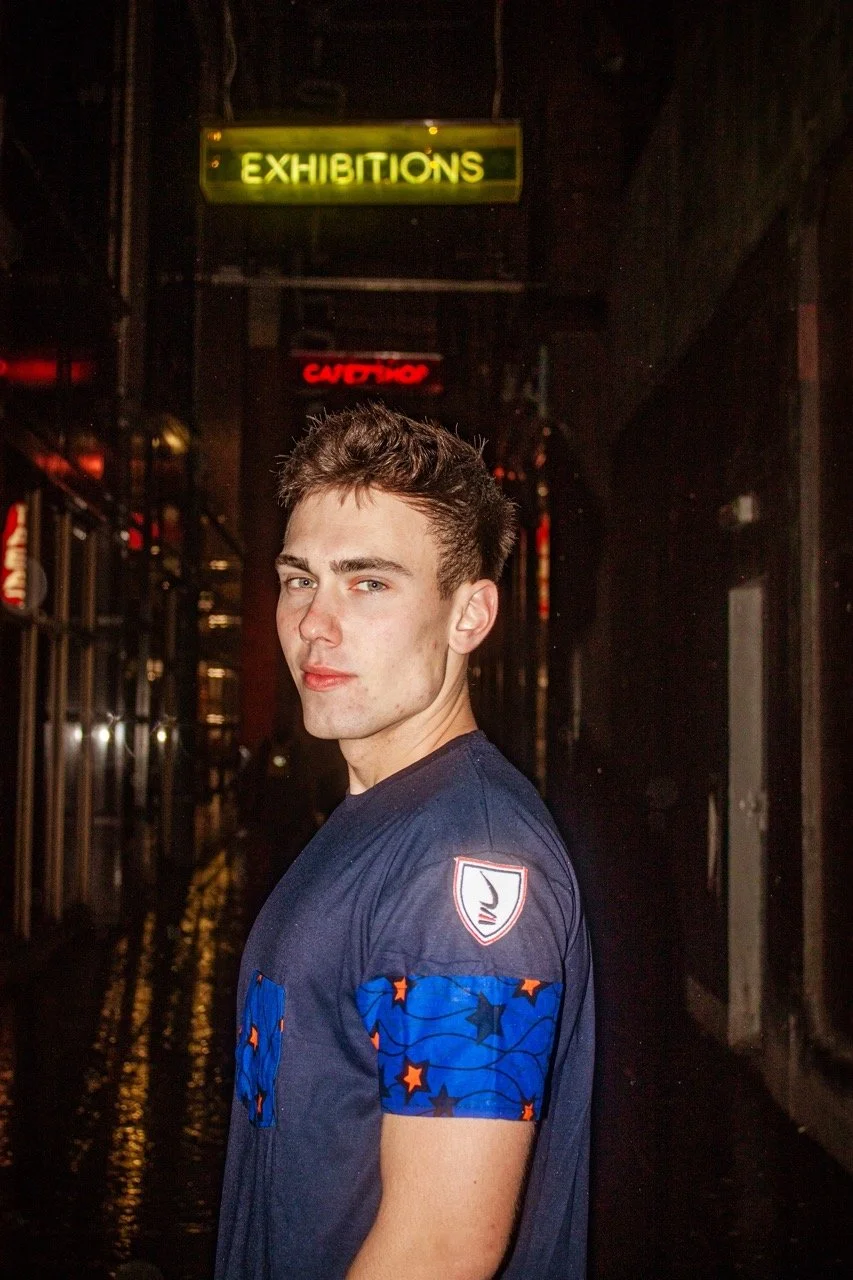 A young man with short messy hair standing in a dimly lit alley, wearing a dark blue sports jersey with patches, with neon signs reading 'EXHIBITIONS' and 'CAUTION' in the background.