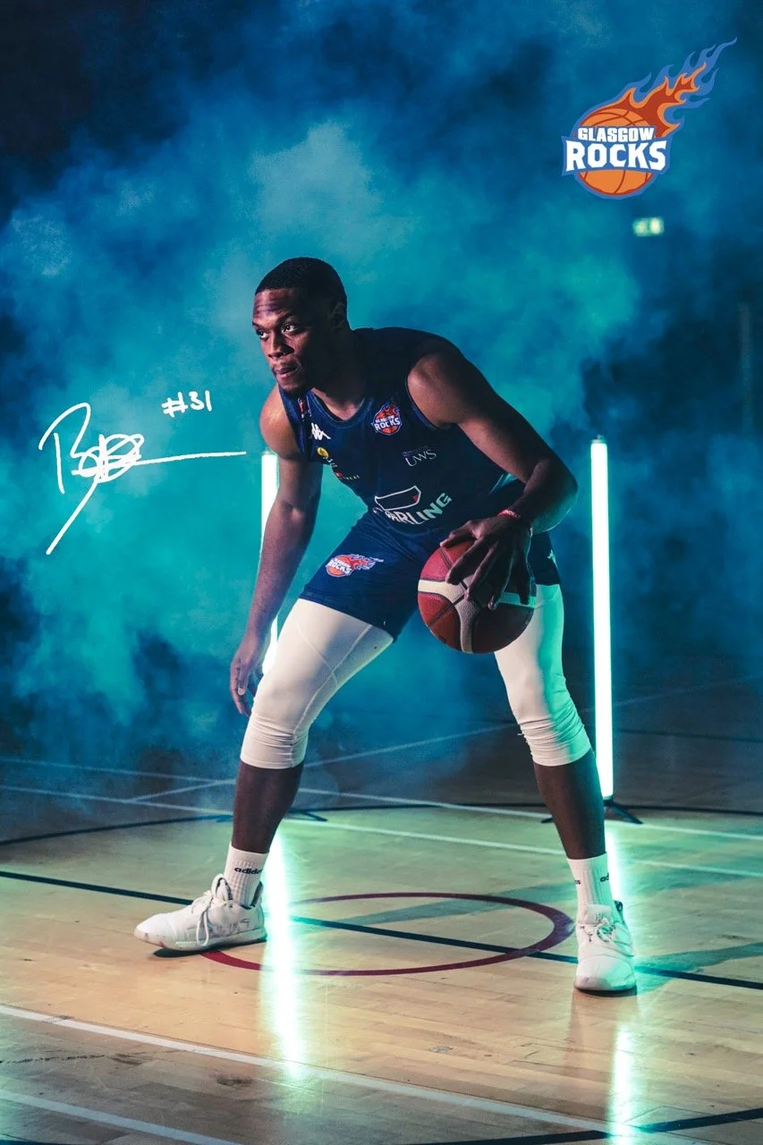 Basketball player in a gliding stance holding a basketball on a court with neon lights and Glasgow Rocks team branding, dark smoky background with blue lighting.