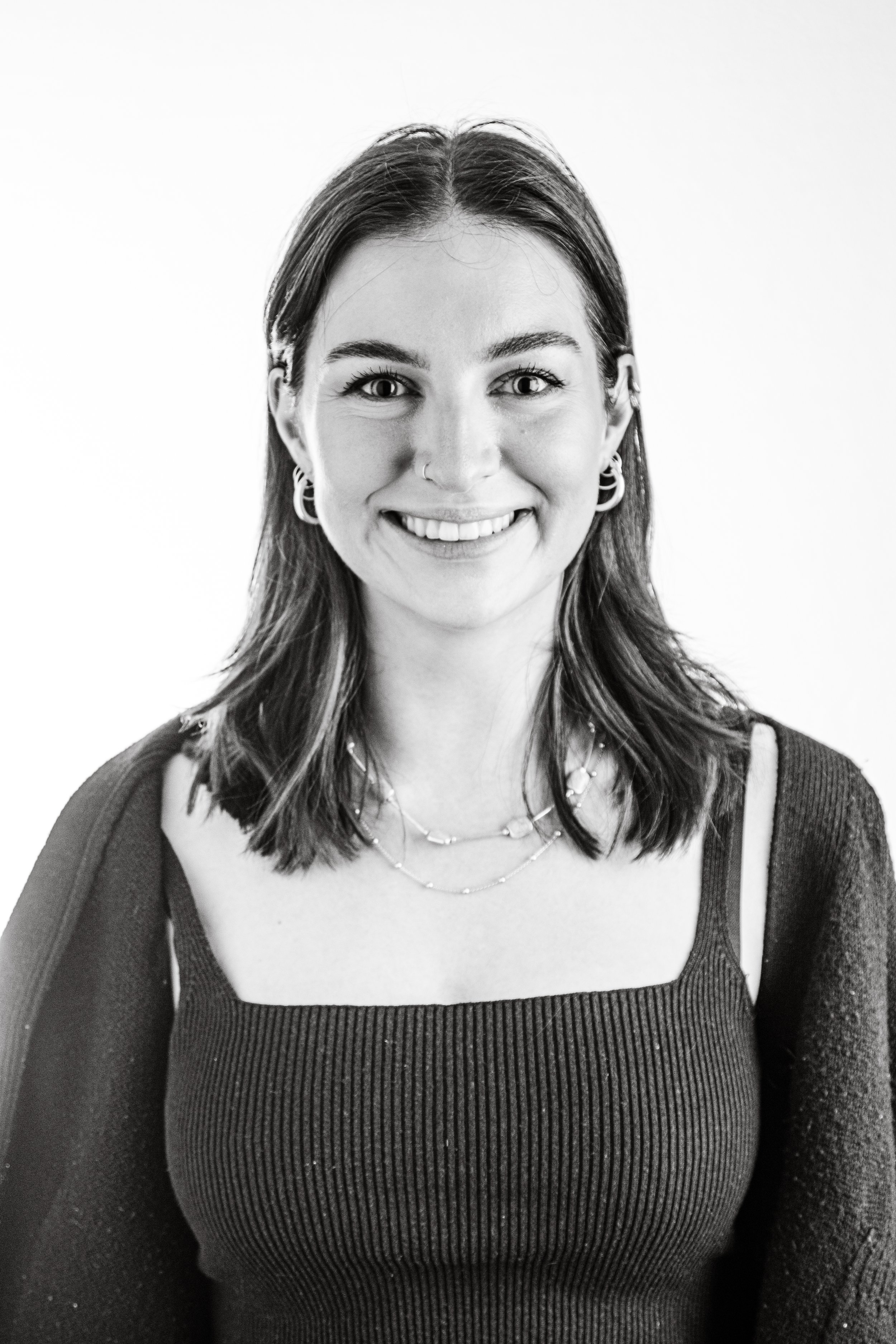Black and white portrait of a smiling woman with shoulder-length hair, wearing layered necklaces, earrings, and a ribbed tank top.