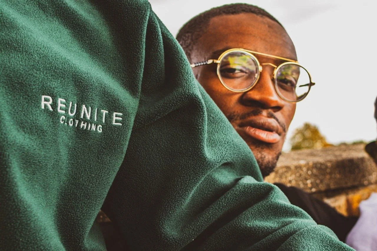 A man wearing glasses and a green hoodie with 'REUNITE CLOTHING' embroidered on the sleeve, looking over his shoulder outdoors.