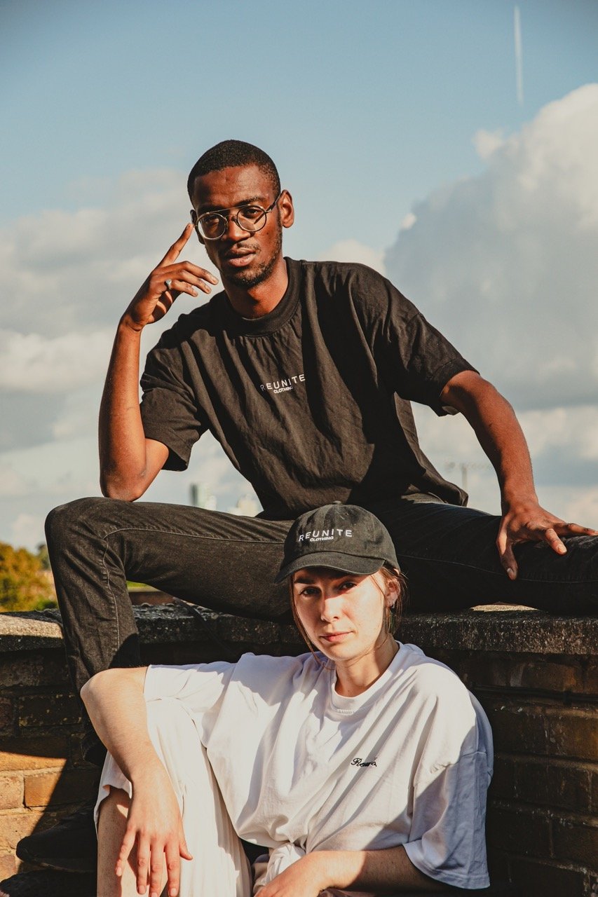 A man and a woman sitting outdoors on a stone structure with a partly cloudy sky behind them. The man, wearing glasses and a dark T-shirt, is sitting with one leg over the other and resting his head on his hand. The woman, wearing a white T-shirt and