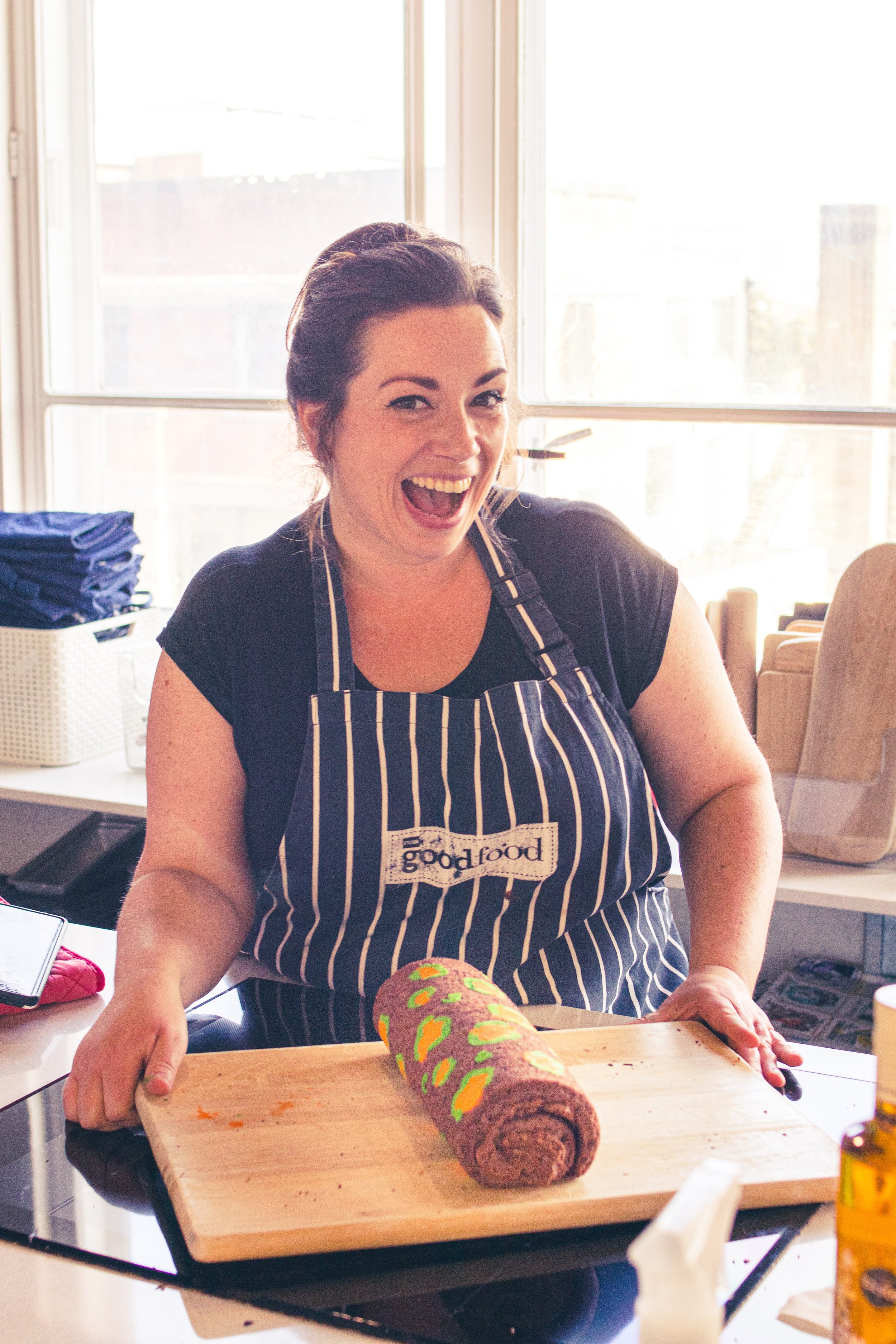 Woman smiling and showing a chocolate roll cake decorated with colorful oval shapes on a wooden cutting board in a kitchen.