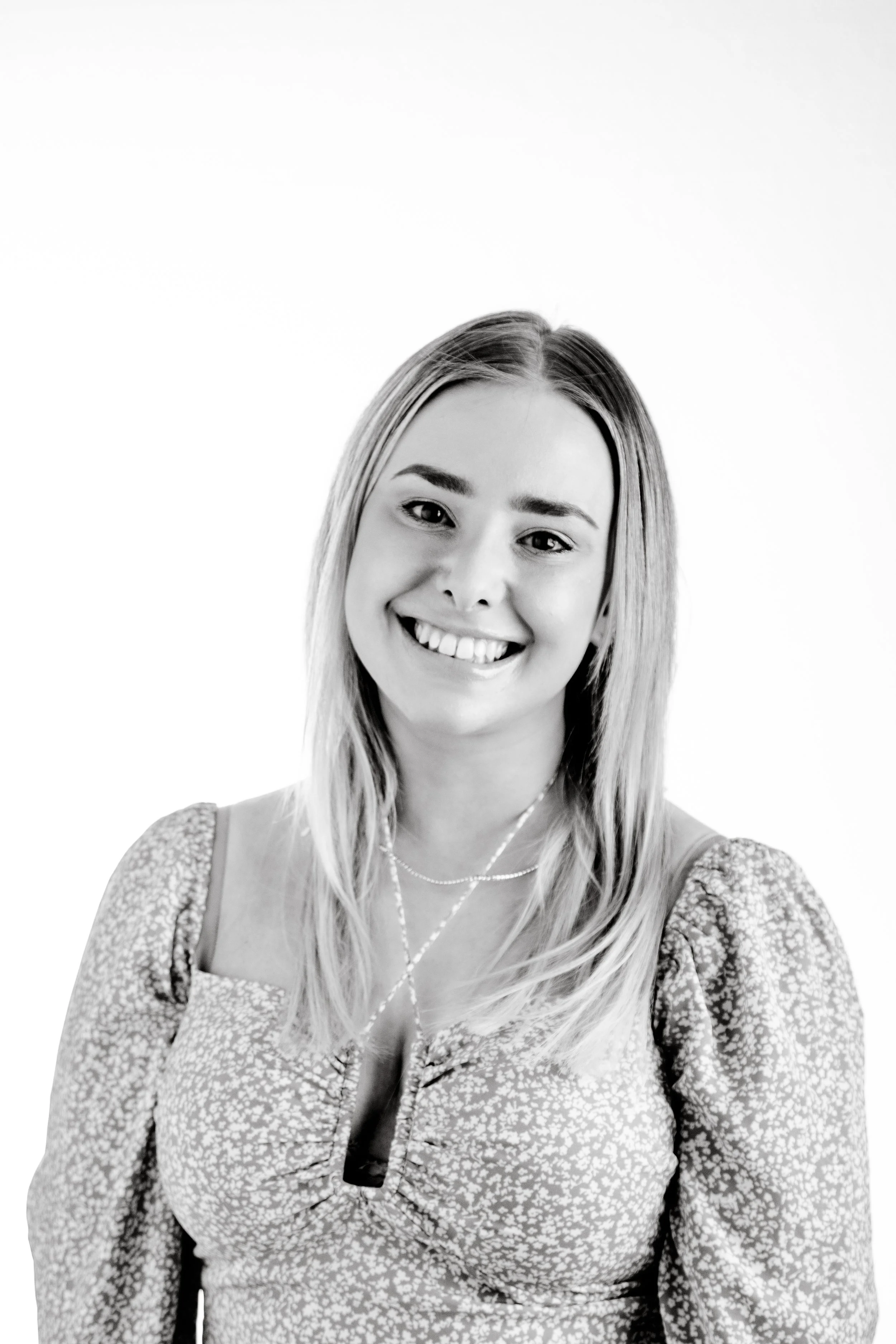 Black and white portrait of a young woman with shoulder-length hair, smiling, wearing a patterned top with puffed sleeves and layered necklaces.