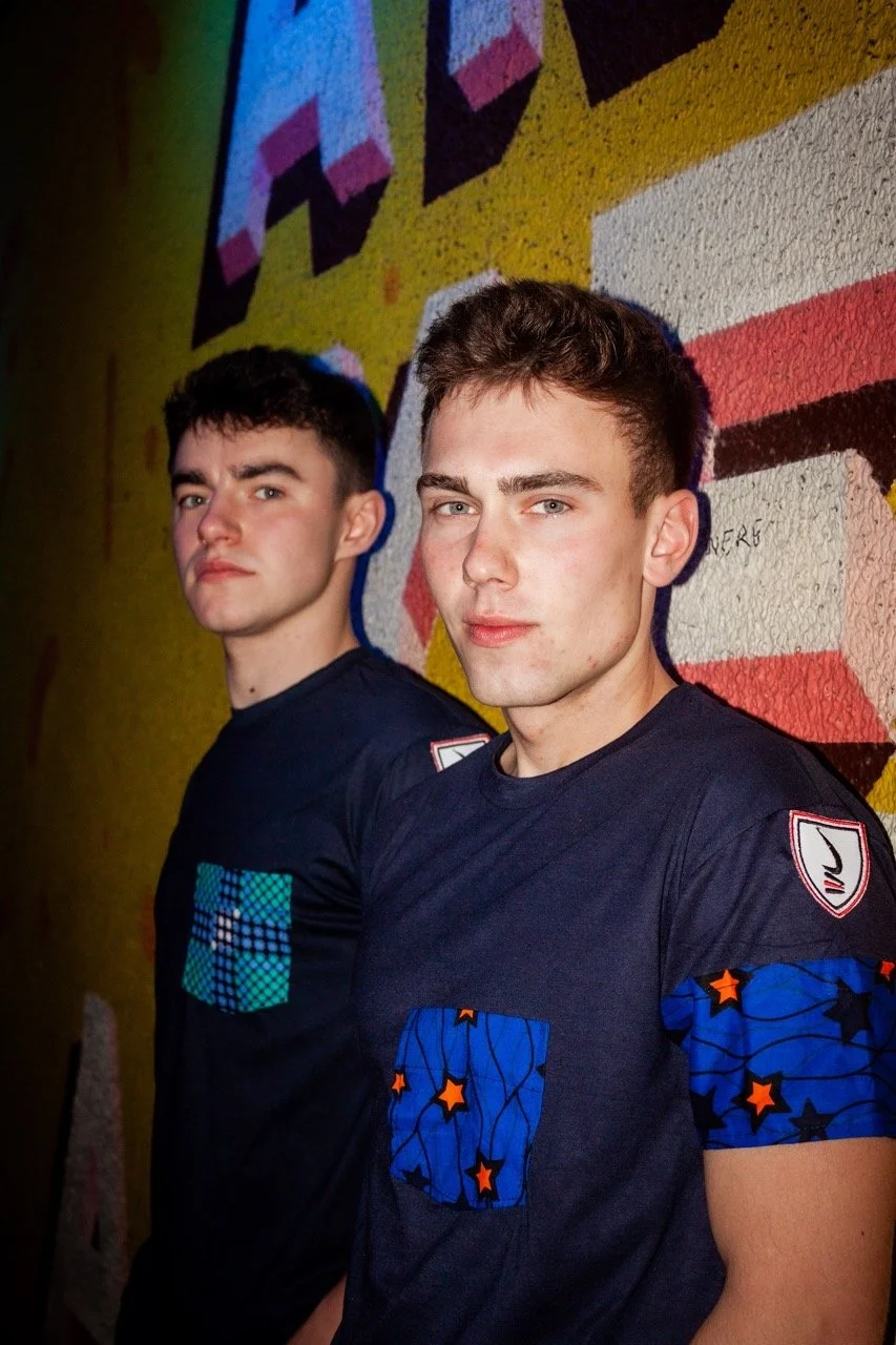 Two young men stand side by side in front of a colorful graffiti wall, wearing black T-shirts with patterned patches on the sleeves and chest.