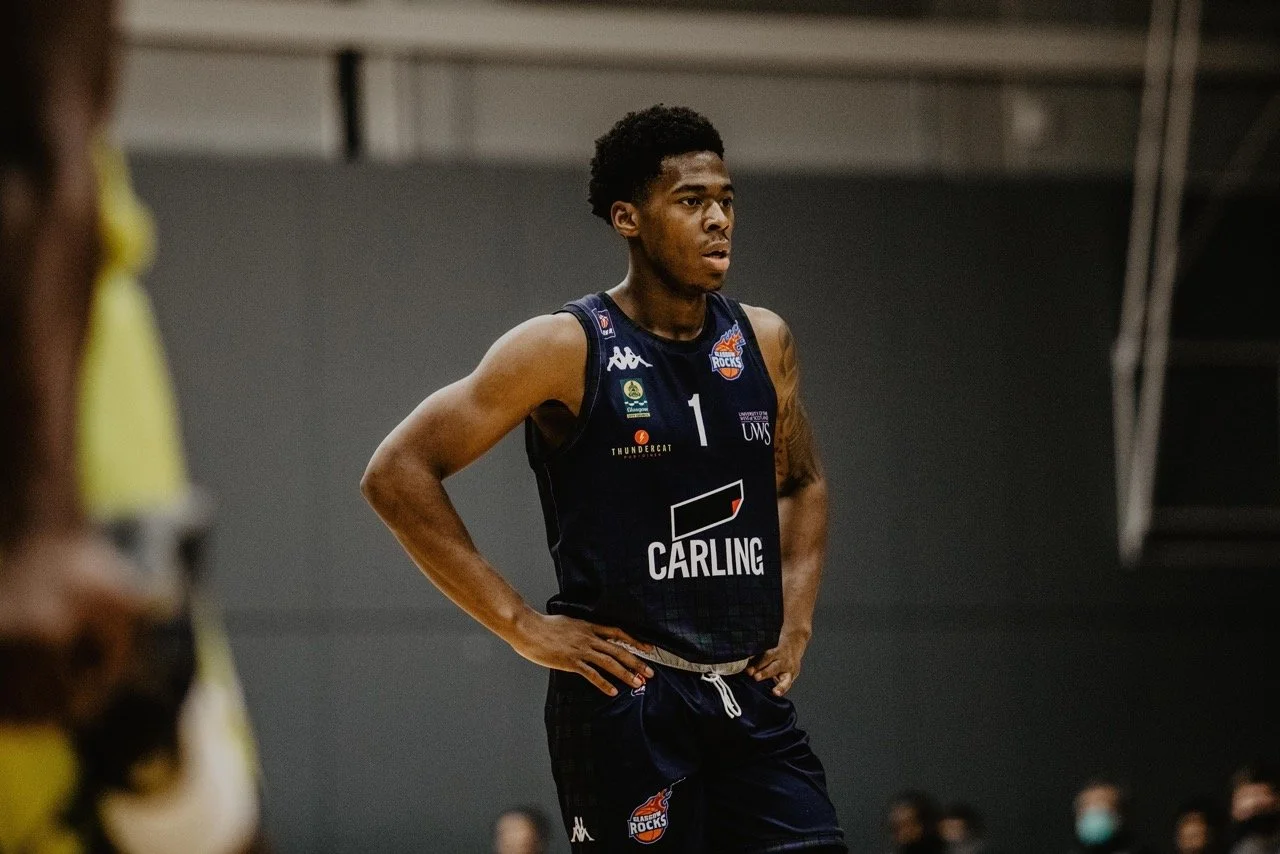 A young male basketball player standing with hands on hips, wearing a dark blue jersey with multiple sponsor logos, during a game or practice.