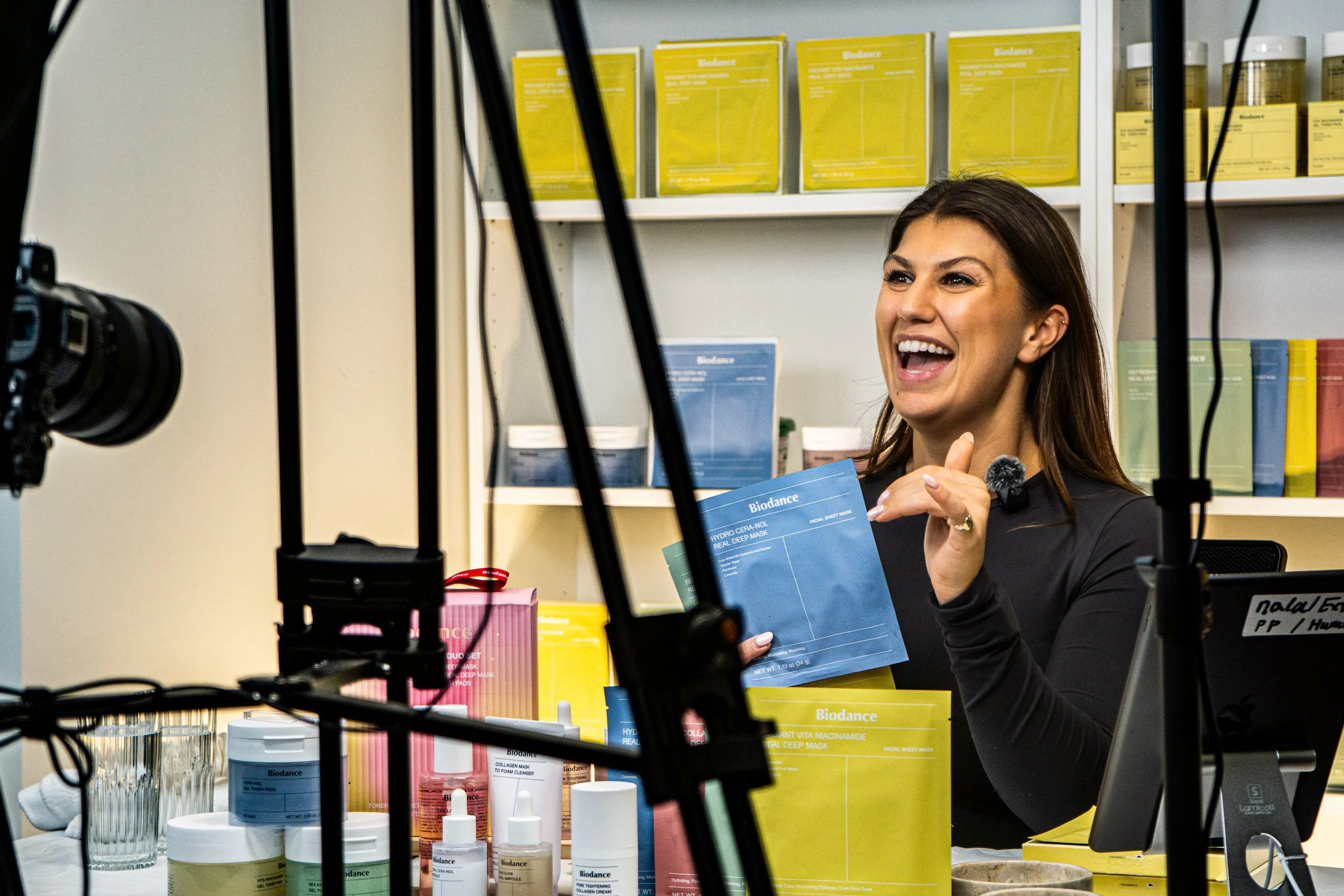 A woman smiling happily at a product review or demonstration in a store or studio, surrounded by skincare or beauty products on the table and shelves.