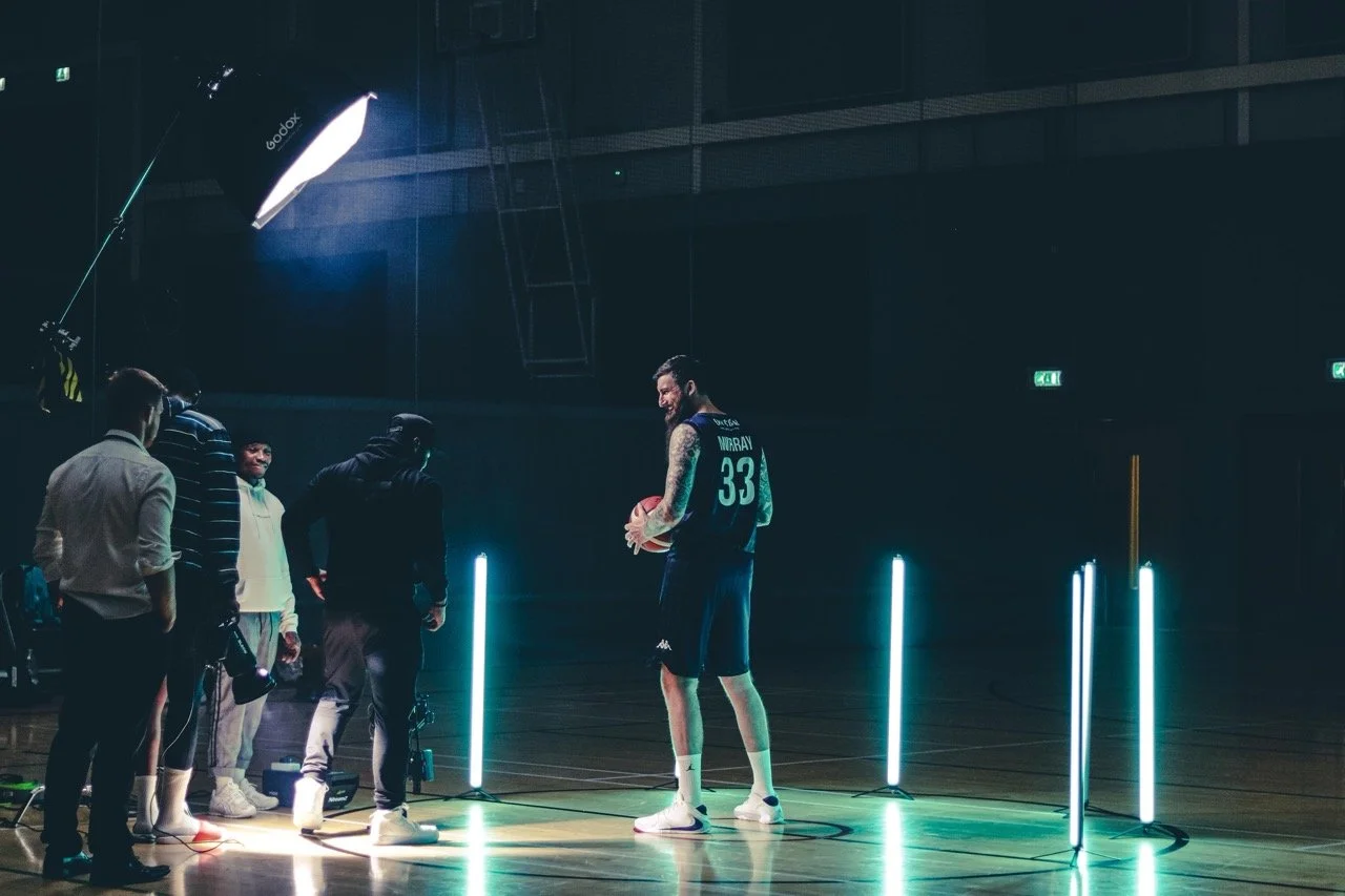 A basketball player with jersey number 33 holding a basketball in a dark gym, surrounded by crew members, with vertical blue and white lights creating a modern atmosphere.
