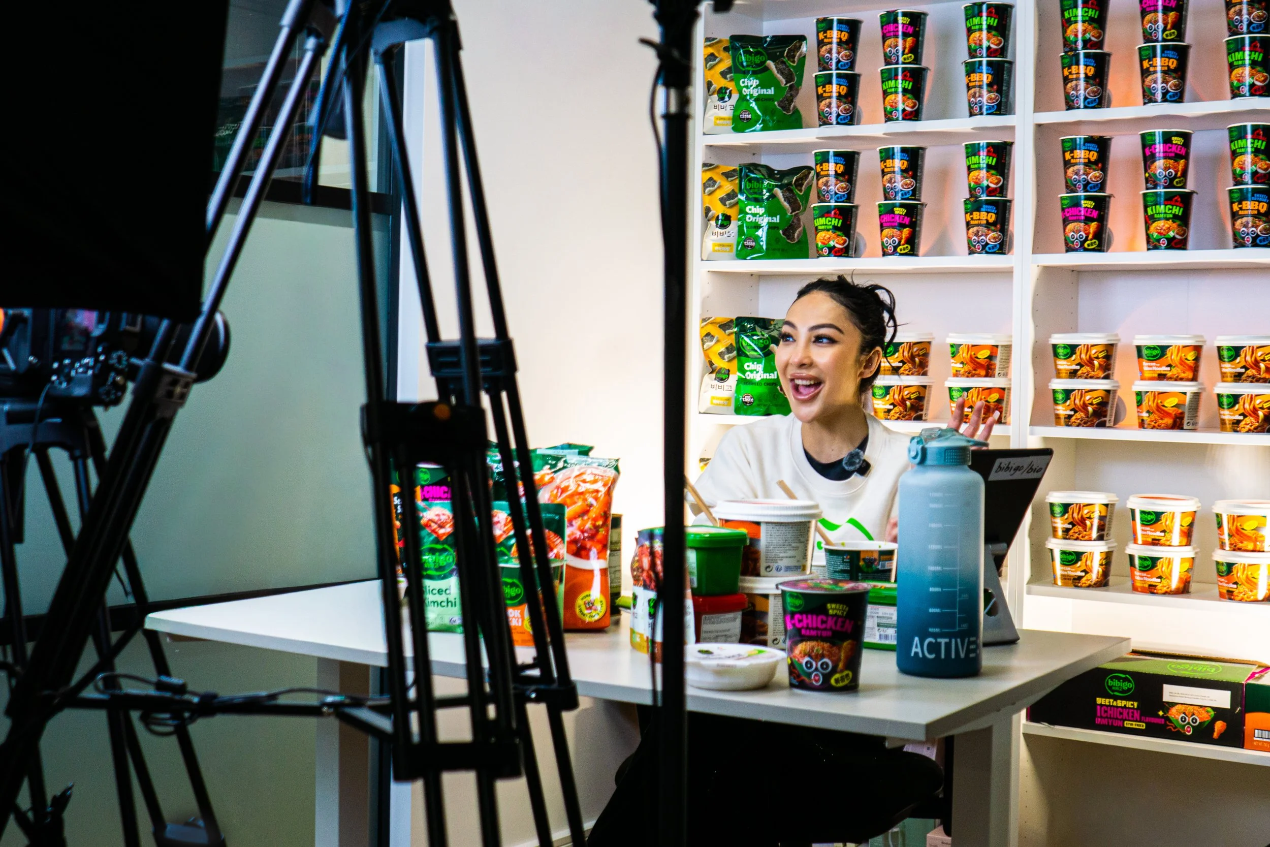 A woman is filming a product review or promotional video for snack foods in front of a shelf stocked with various instant noodle cups and snack packages, inside a store or studio.