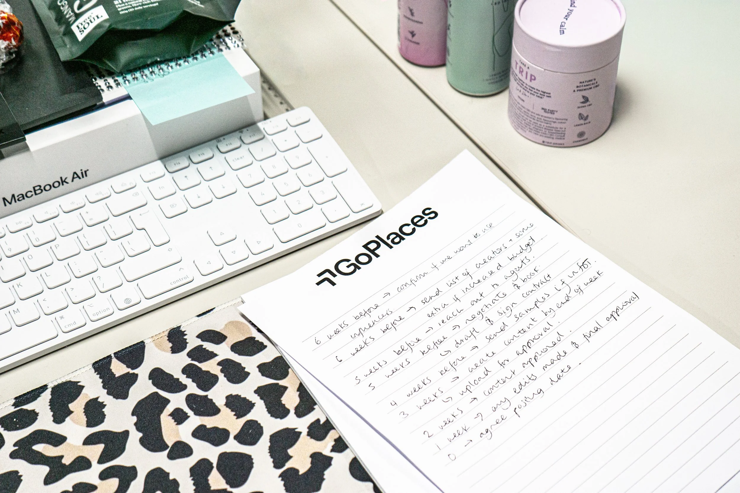 A workspace with a white Apple MacBook Air computer, a white wireless keyboard, a spiral notebook with handwritten notes titled 'Go Places', a leopard print notebook, and several cans of Trippy brand beverages on a beige desk.