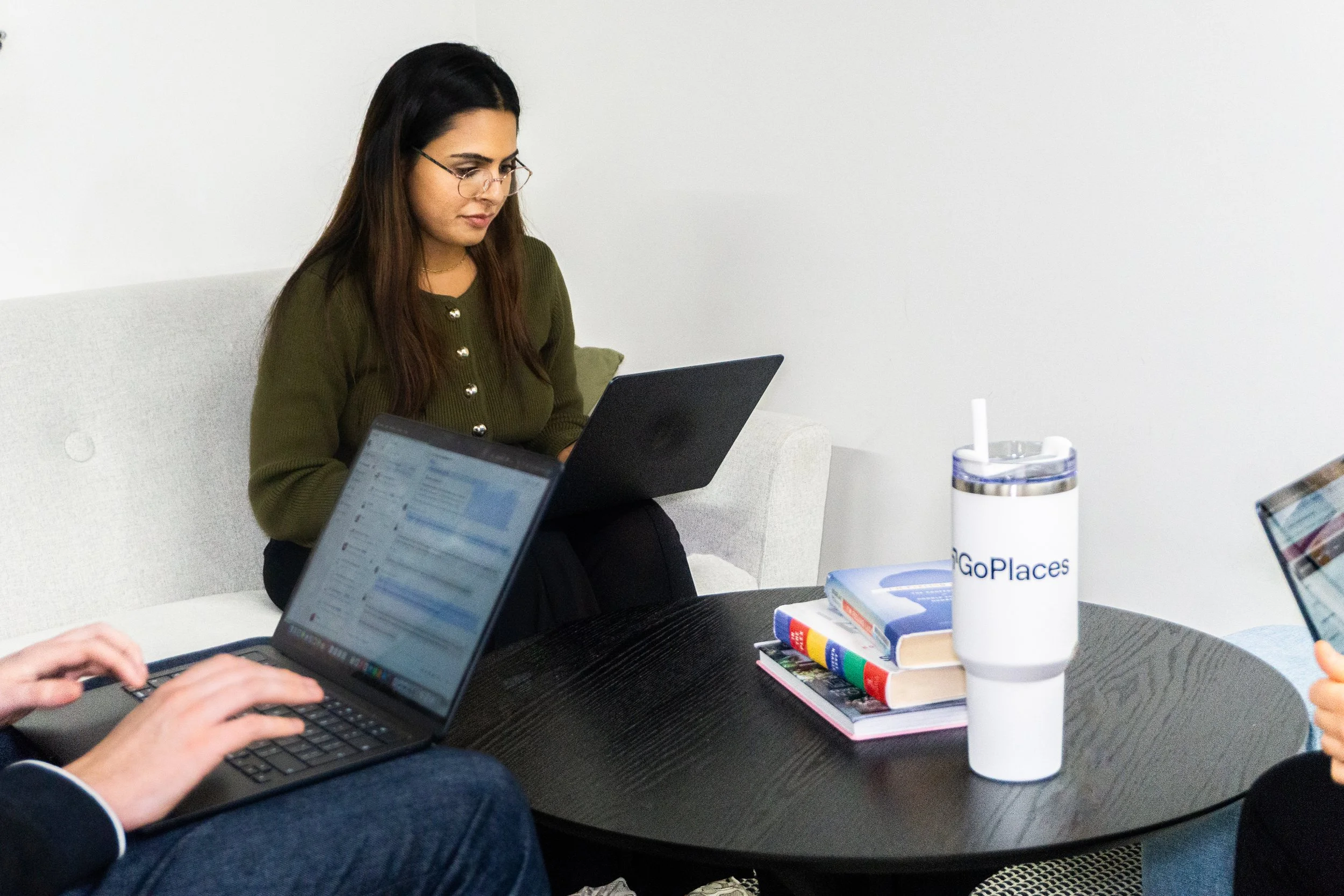 Three people with laptops sitting around a round black table in a room with white walls, one woman in a green sweater with glasses focused on her laptop, and books and a white insulated cup with a straw on the table.