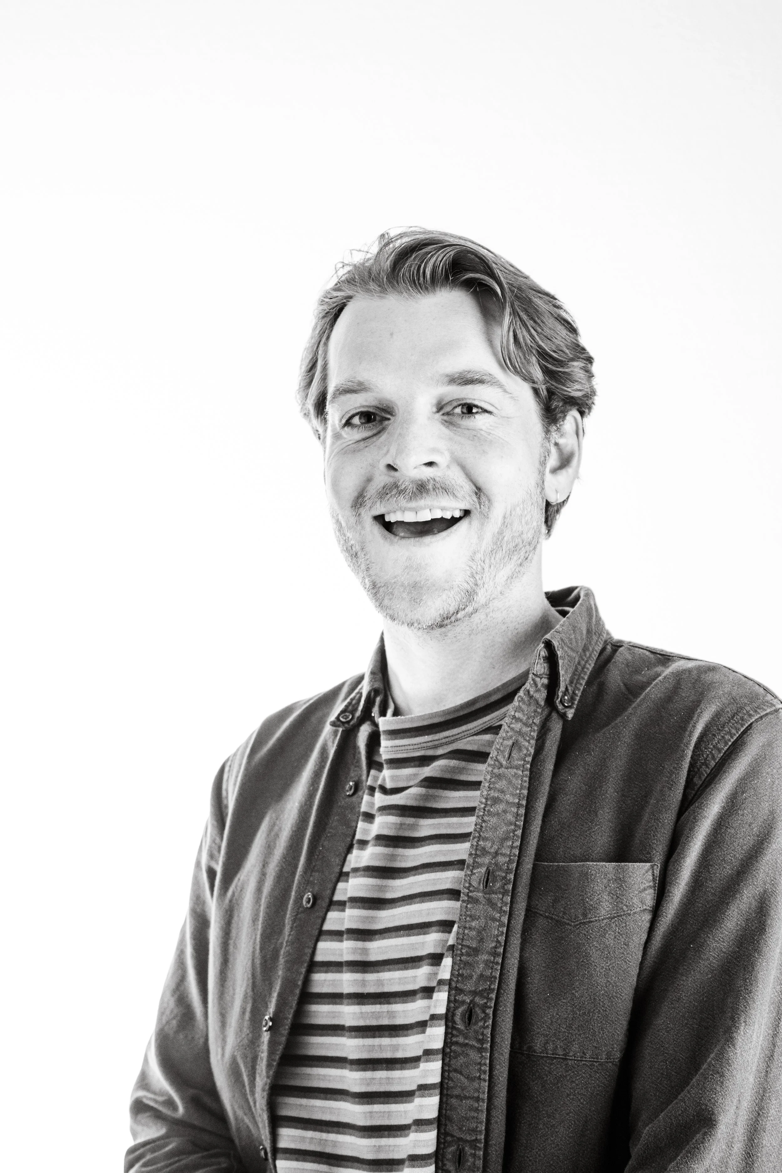 A black and white portrait of a smiling man with medium-length hair, wearing a striped t-shirt and an unbuttoned shirt over it, against a plain light background.