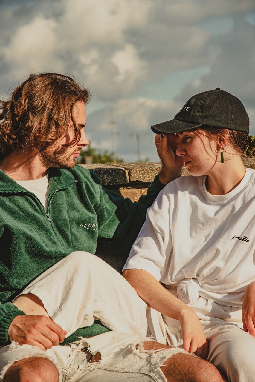 A young man with long curly hair and a beard, wearing a green fleece jacket, and a young woman with reddish hair, wearing a black cap and a white T-shirt, sit closely together outdoors. The man touches the woman's forehead gently, and they appear to 