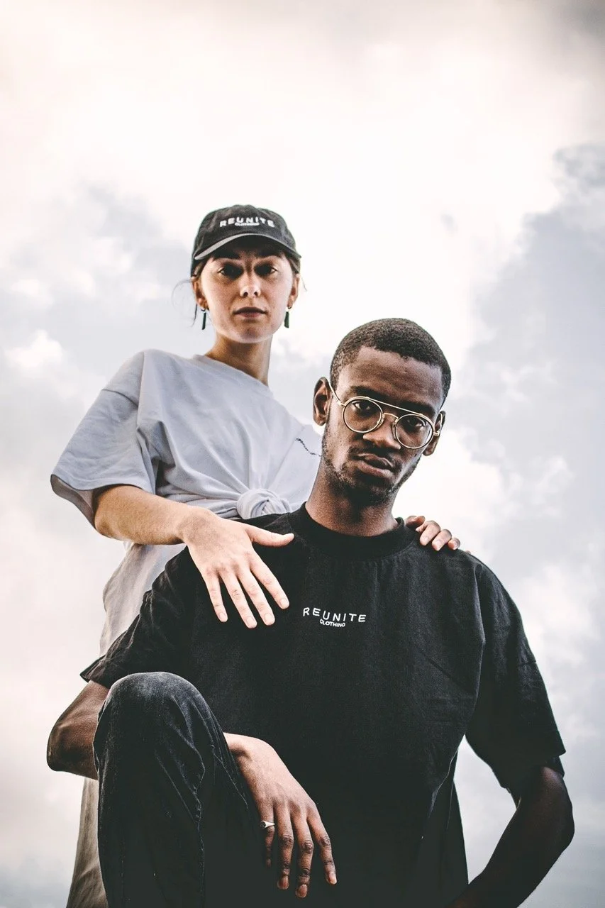 A woman standing behind a man, both looking serious, outdoors under cloudy sky. The woman has a black cap and light-colored shirt, the man wears glasses and a black shirt with 'REUNITE' written on it.