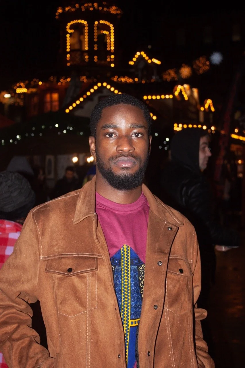 A young man with dark hair and beard wearing a brown jacket and pink shirt stands outdoors at night, with festive lights and a carnival ride in the background.