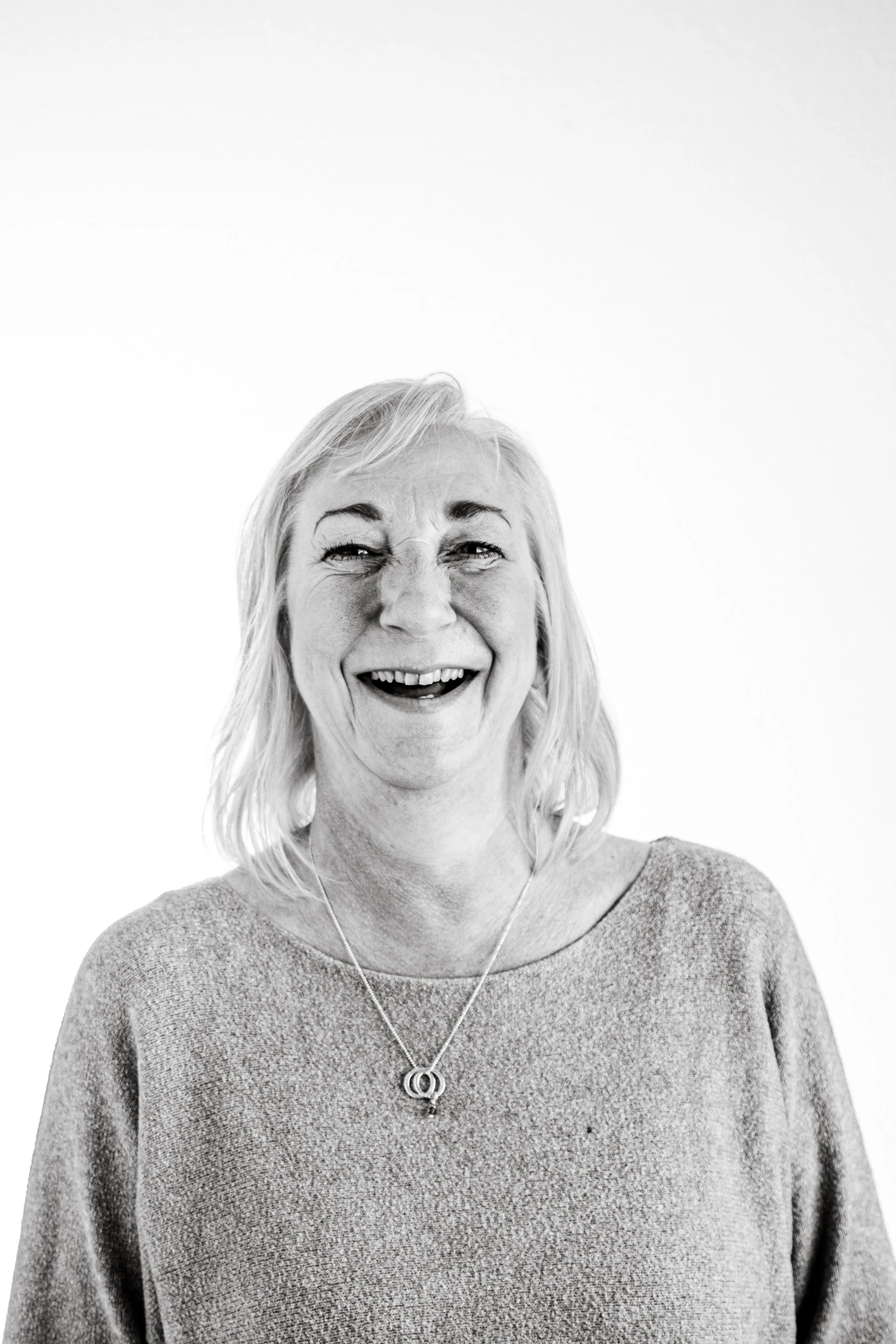 A black-and-white photo of a smiling woman with shoulder-length light hair, wearing a round-neck sweater and a necklace, standing against a plain white background.