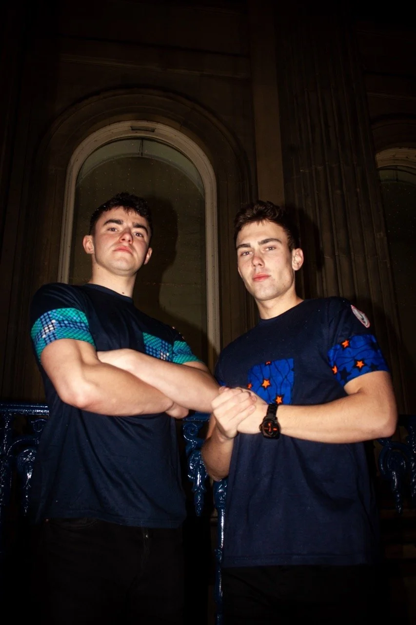Two young men stand in a dimly lit room, facing forward with serious expressions. They are gripping each other's hands in a handshake or arm wrestling pose. The background features large arched windows and dark wood paneling.