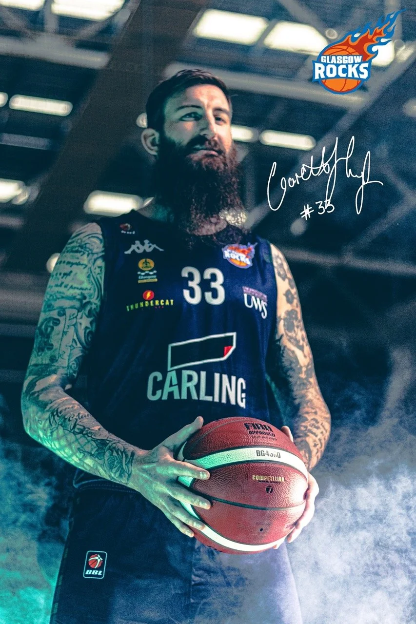 A male basketball player with tattoos on his arms, wearing a navy blue jersey with the number 33, holding a basketball, and standing under a sports arena ceiling. There are logos for Glasgow Rocks, Carling, and other sponsors on his jersey, and a han