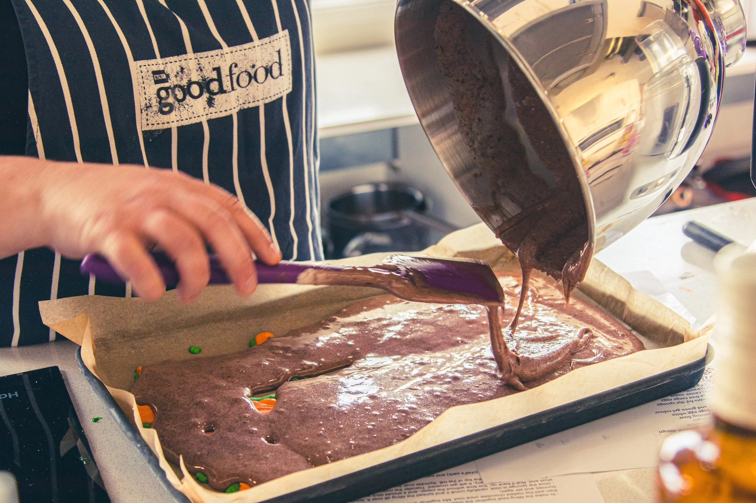 Person pouring chocolate batter onto green and orange candy in a baking tray, wearing a striped blue and white apron.