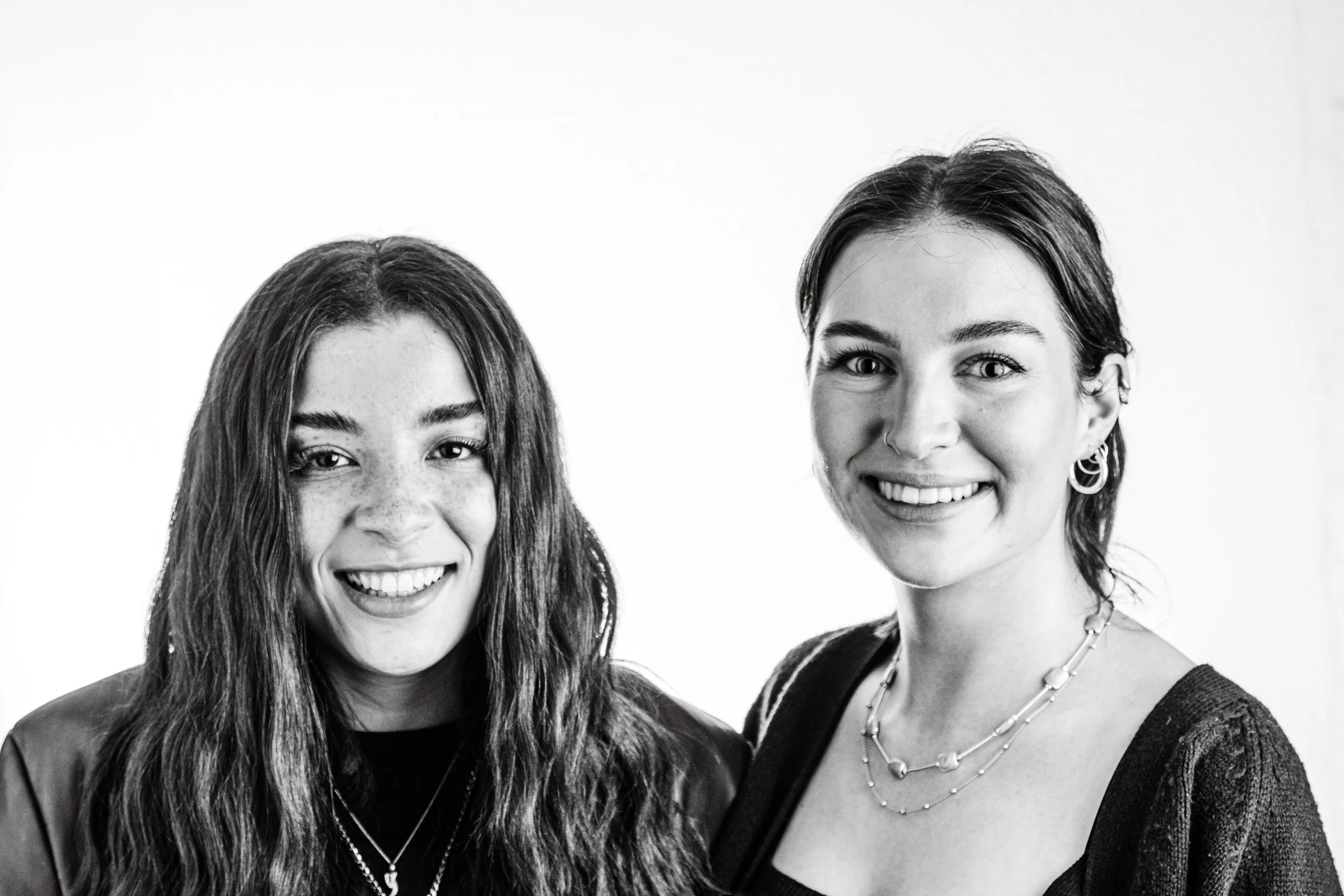 Two young women smiling, one with long wavy hair and freckled face, the other with short dark hair and earrings, in black and white.