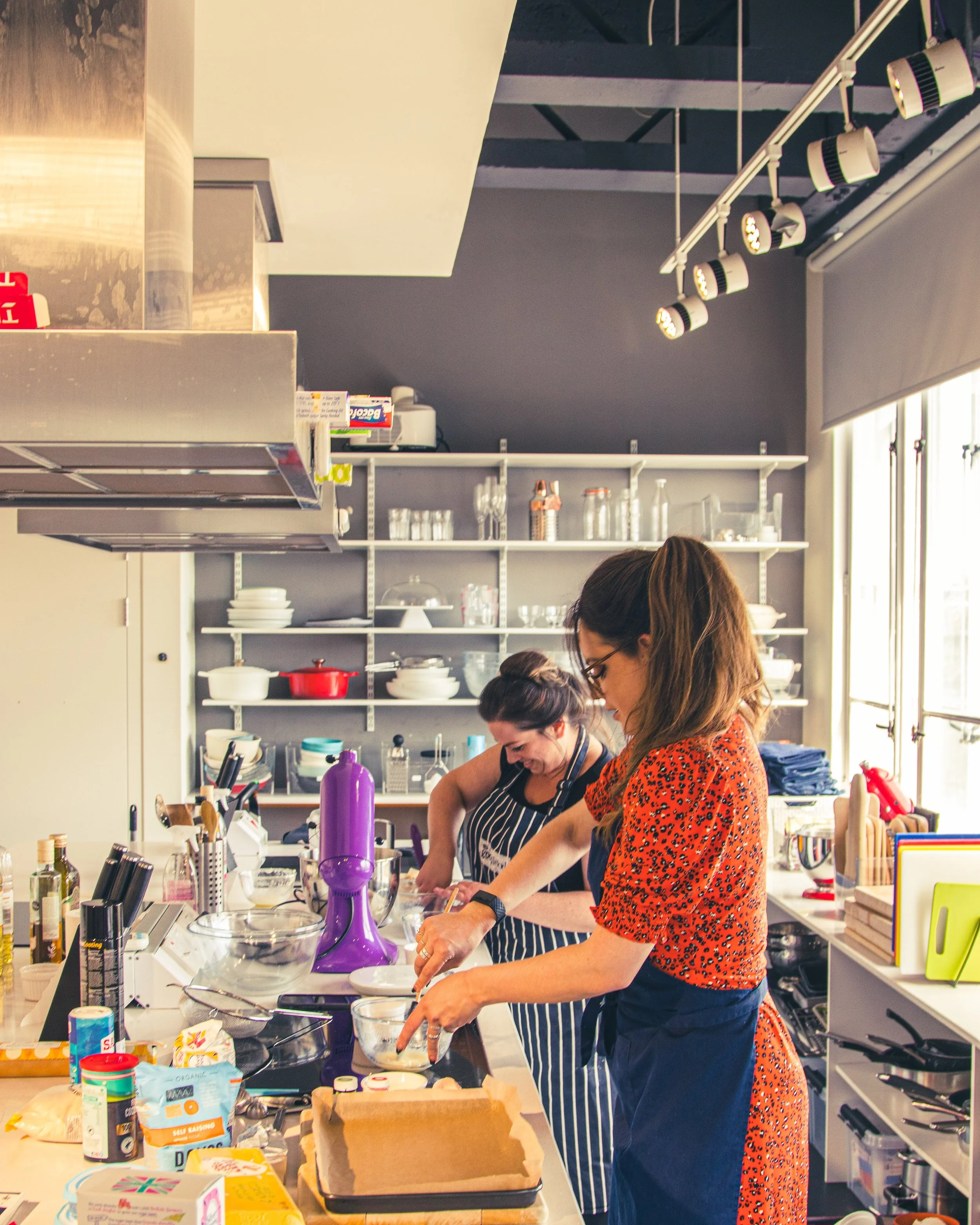Two women baking together in a modern kitchen, preparing food on a large countertop surrounded by baking ingredients and utensils.