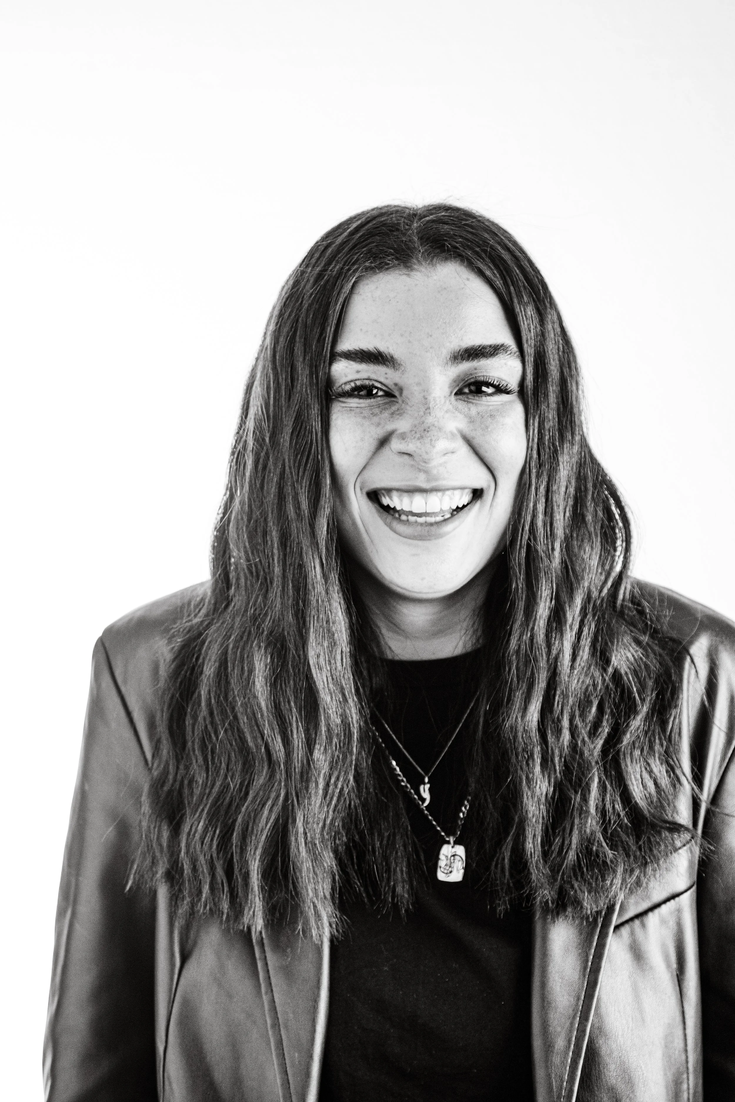 Black and white photo of a woman with long hair, smiling, wearing a leather jacket and layered necklaces.
