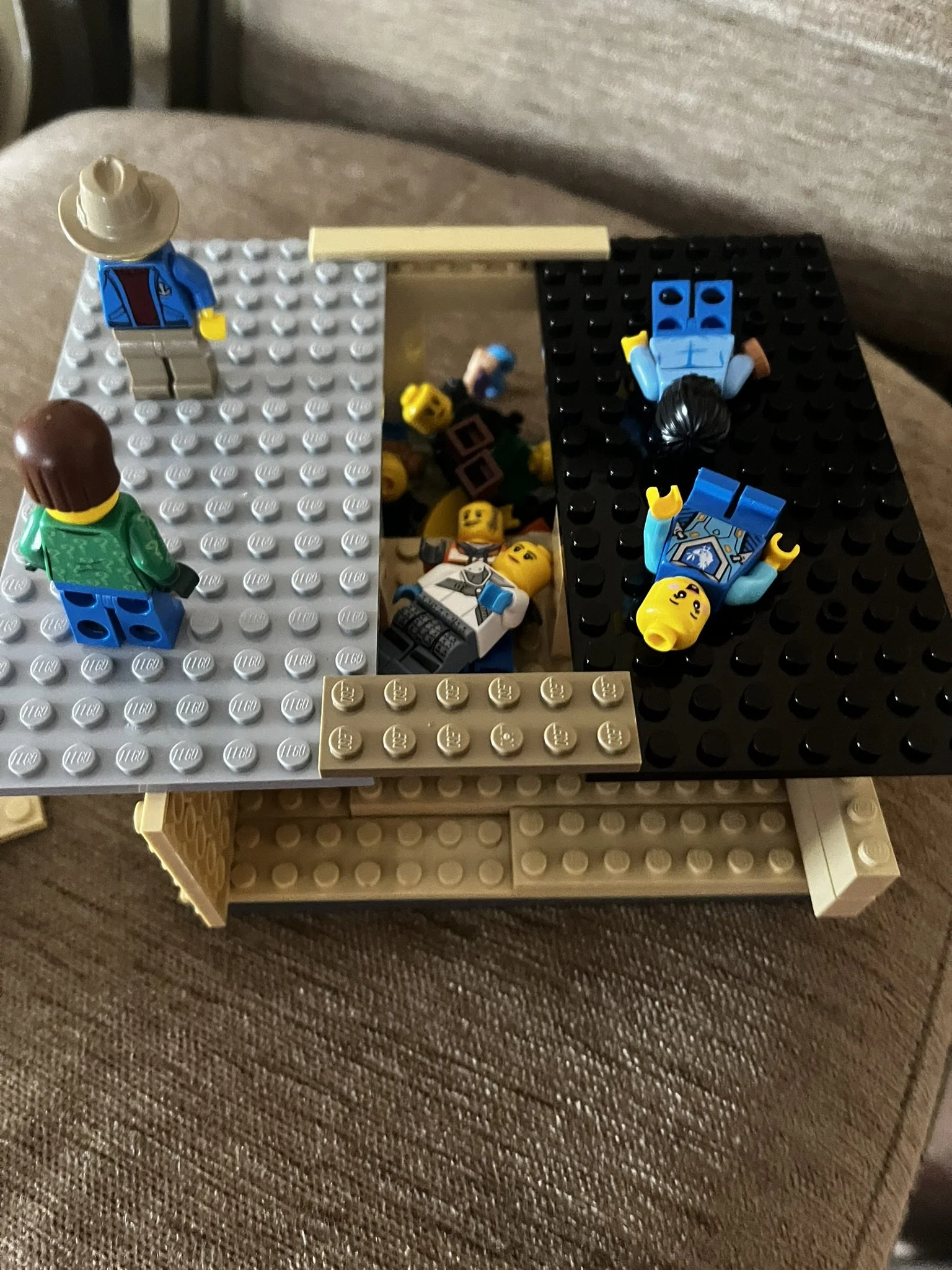 Lego scene with minifigures including a woman with black hair, a man in a white shirt, and a man with brown hair, spread out on a black, gray, and tan baseplate. Some minifigures are lying down, while others stand. The scene is set on a brown texture