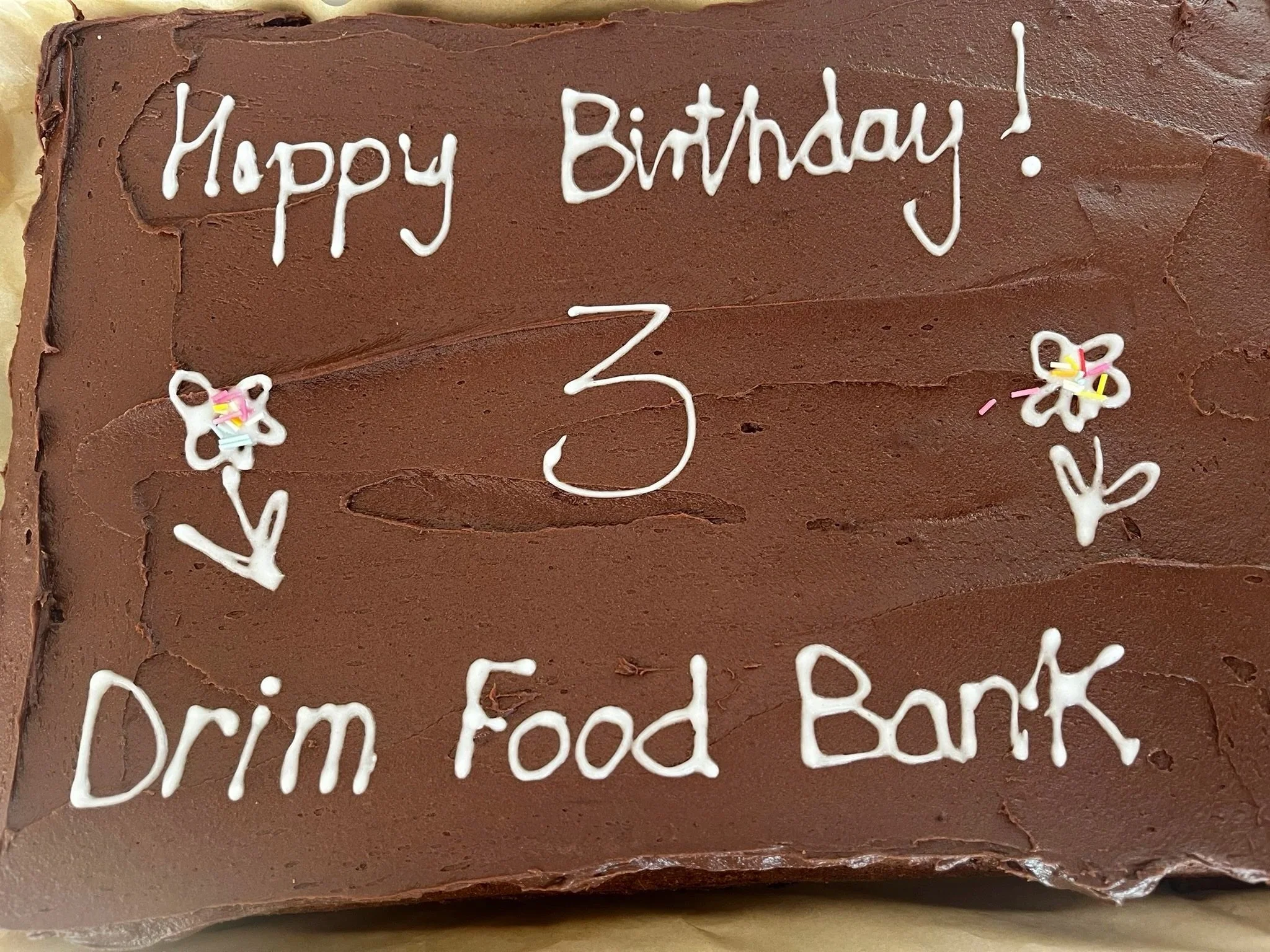 Chocolate birthday cake with white icing writing that says "Happy Birthday!" and "Drim Food Bank". The cake has some flower decorations with pink, yellow, and white sprinkles.