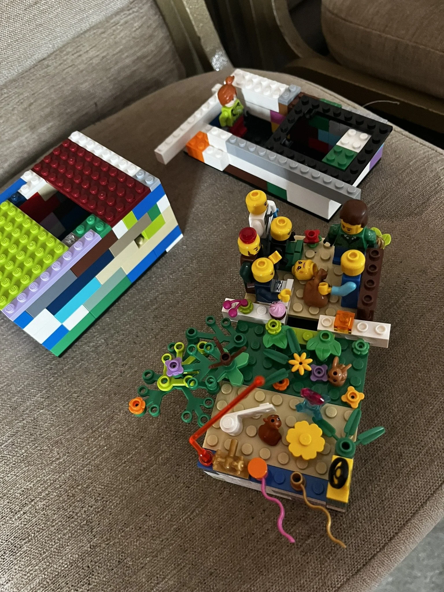 Collection of LEGO sets and minifigures on a brown cushioned surface, including a garden scene with flowers, animals, and tools, and two partially built structures.