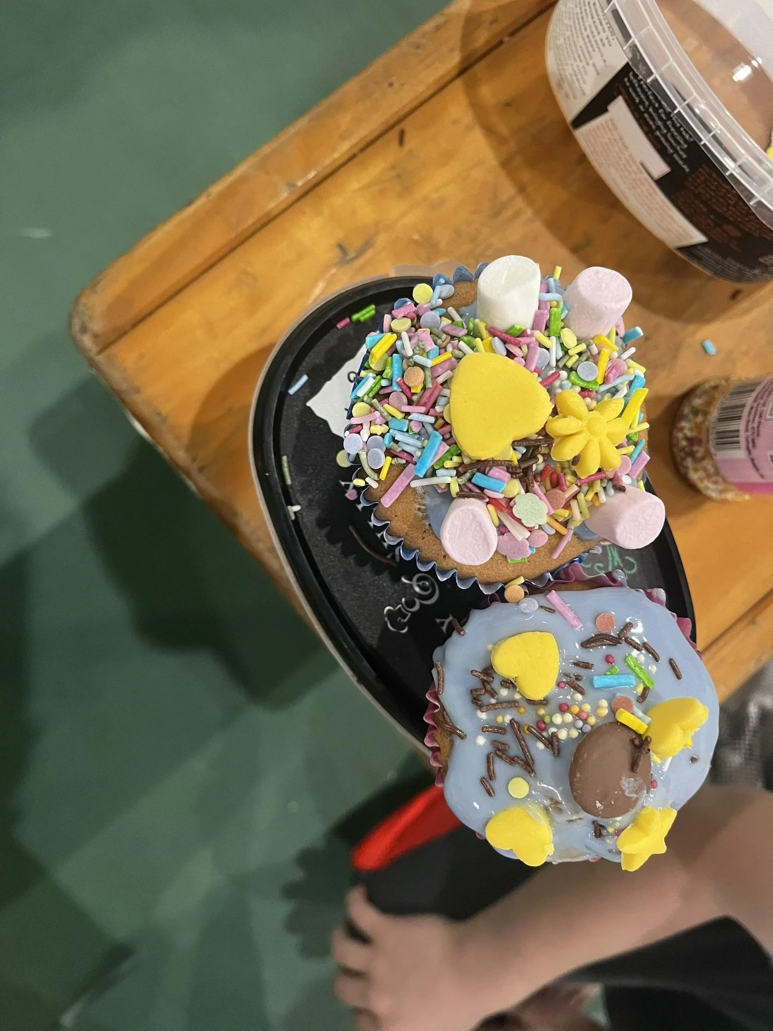 Two cupcakes decorated with colorful sprinkles, candies, and frosting, placed on a black circular tray. One cupcake has blue frosting, and the other has light brown frosting, both topped with small candy hearts and other decorative sweets.