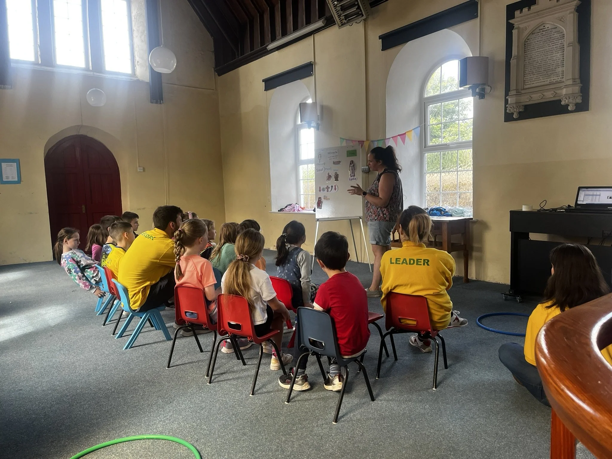 A classroom with a group of children sitting on red and blue chairs, attentively watching a woman standing next to a whiteboard. The woman appears to be teaching or presenting. The room has large windows, and yellow walls with some decorations and a 