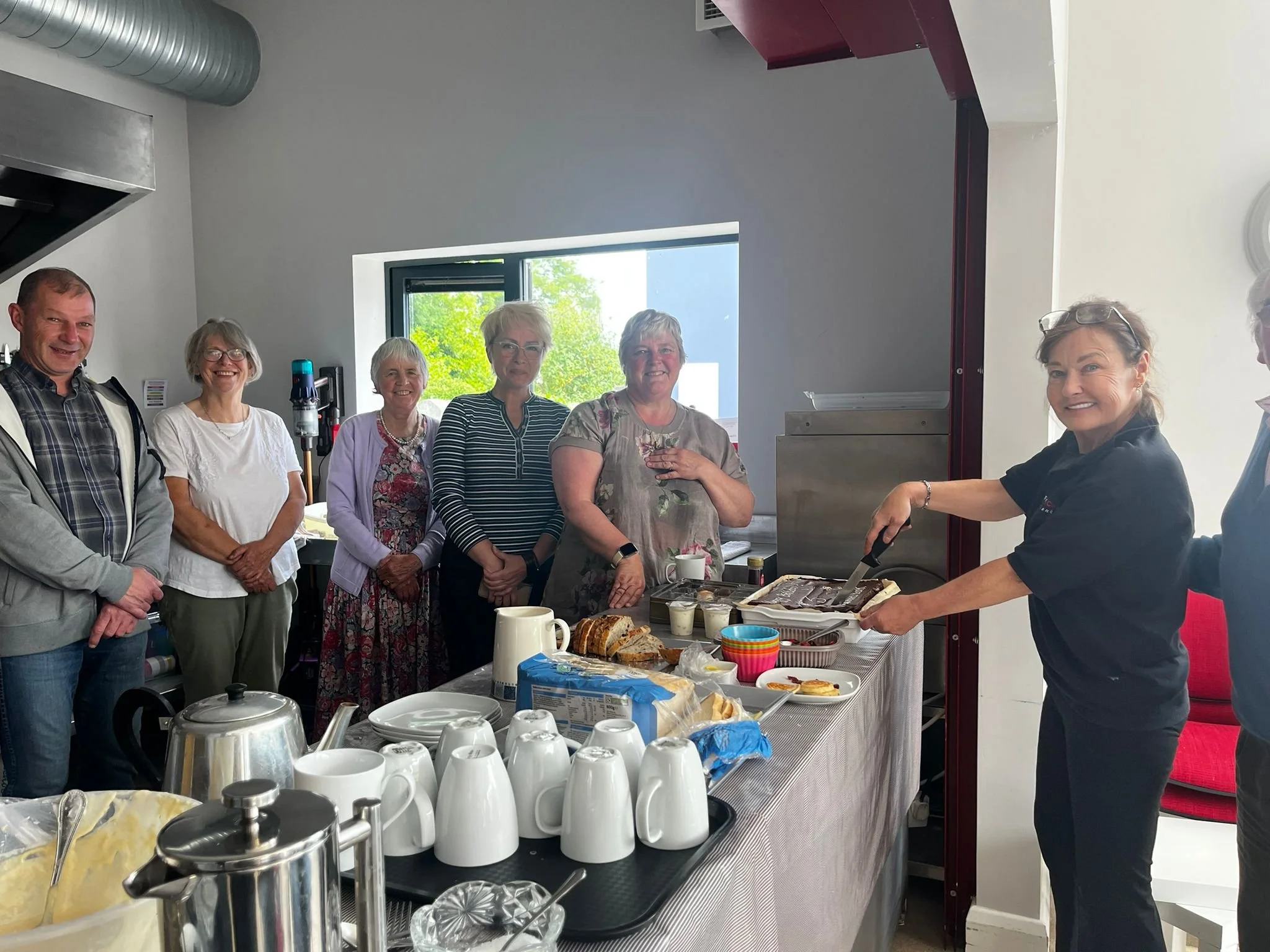 A group of six women and one man standing in a kitchen, with a woman slicing a cake on a table. The table has cups, plates, and food items, and the group is smiling and looking at the camera.