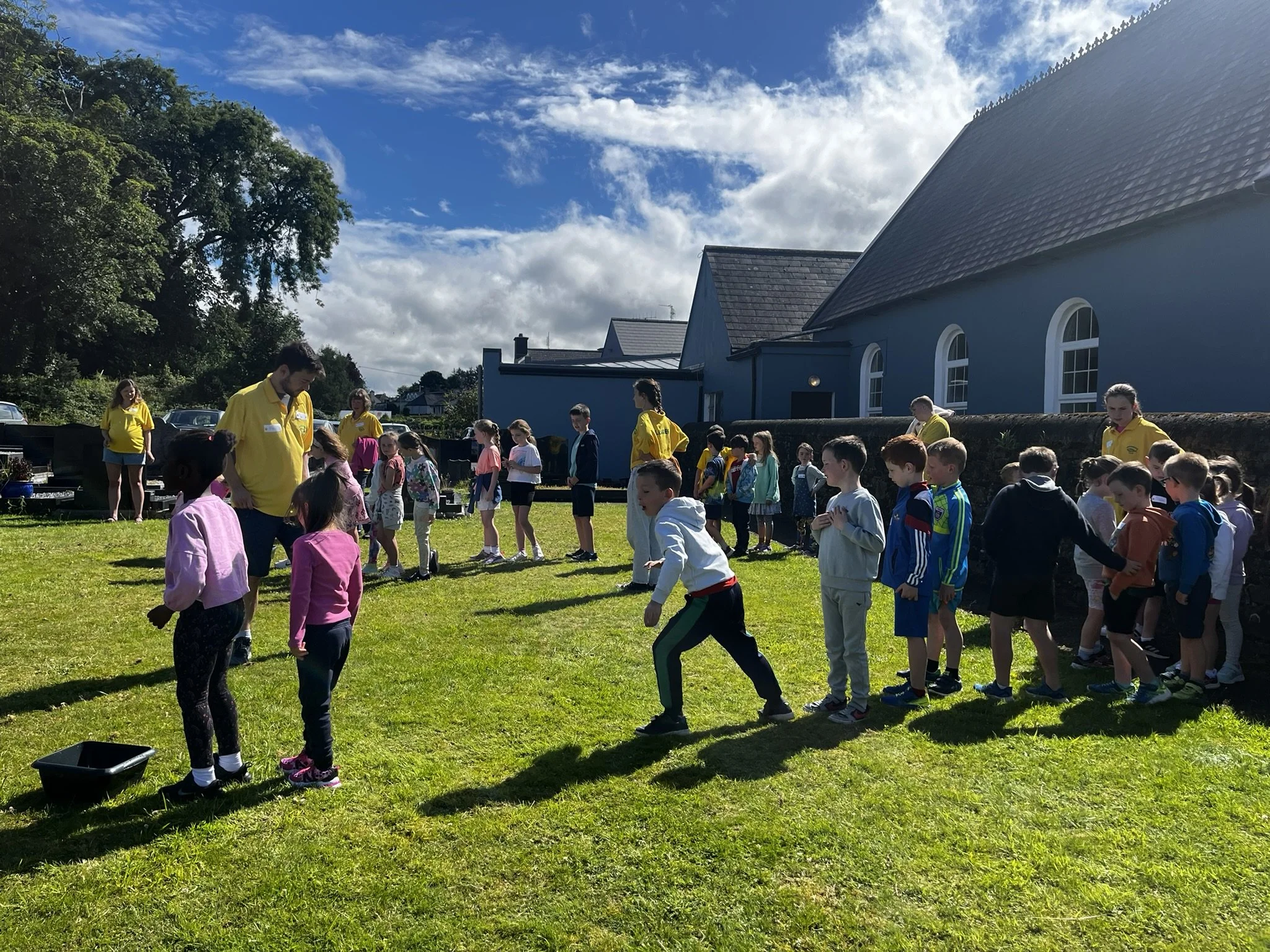 Group of children and adults outdoors on a grassy field, participating in a line game, with blue building and trees in the background under a partly cloudy sky.