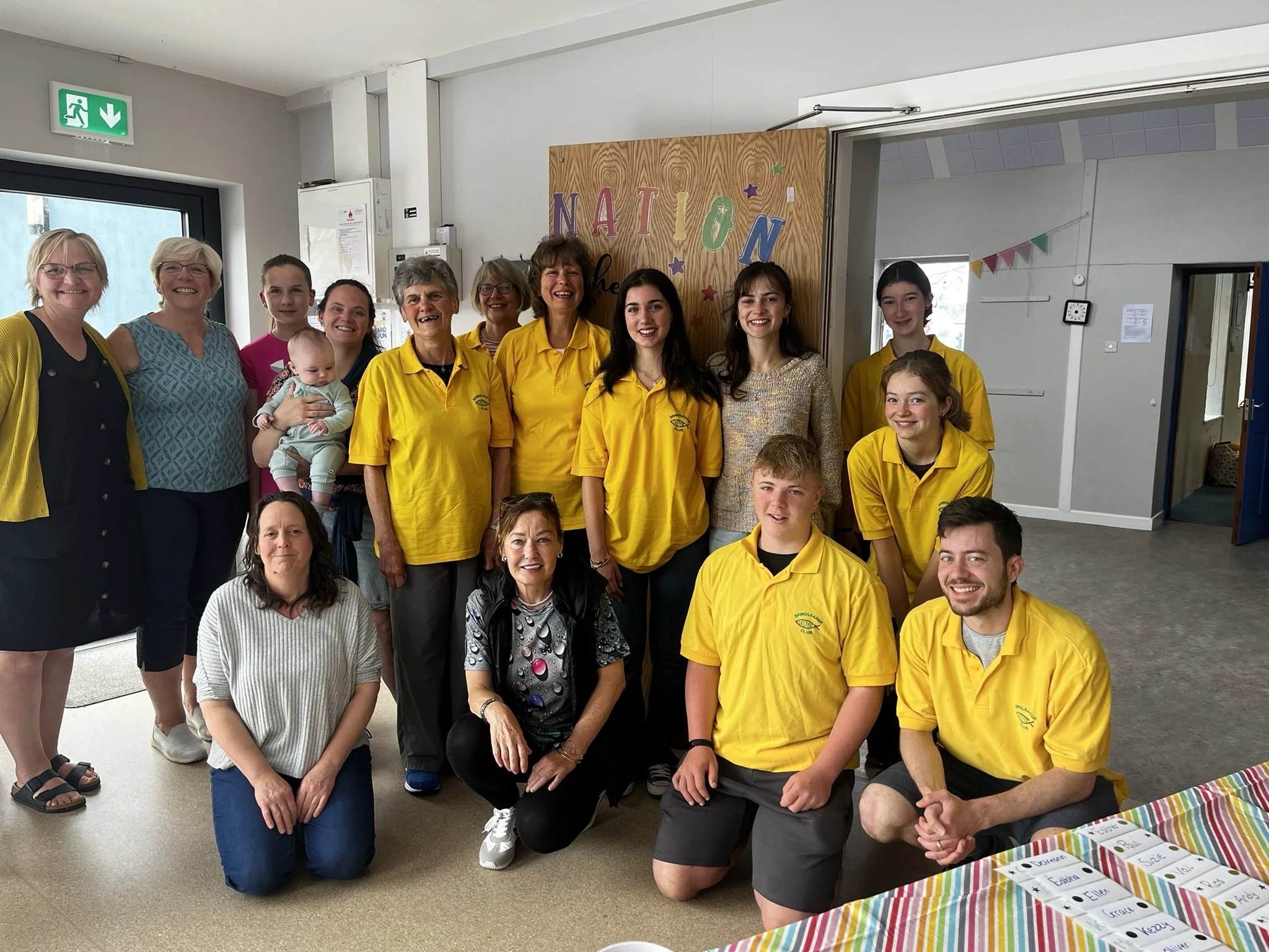 Group of people, including adults and teenagers, posing for a photo indoors in front of a colorful sign that says 'NATION'. Eight individuals are wearing yellow shirts, and they appear to be part of a team or group, with some kneeling or sitting in f