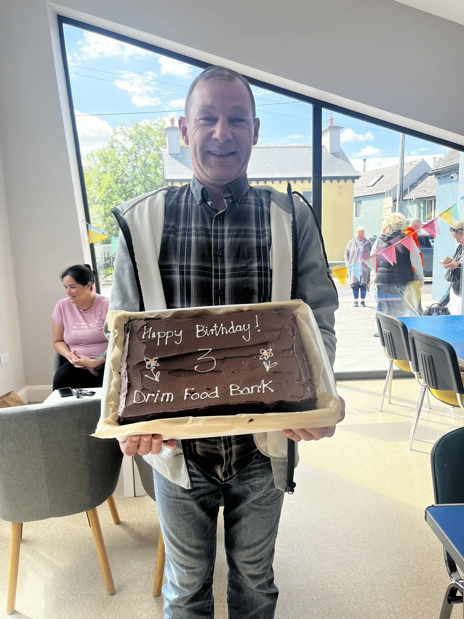 Man holding a birthday cake with a message written in icing, inside a room with large windows showing people outside at a celebration.