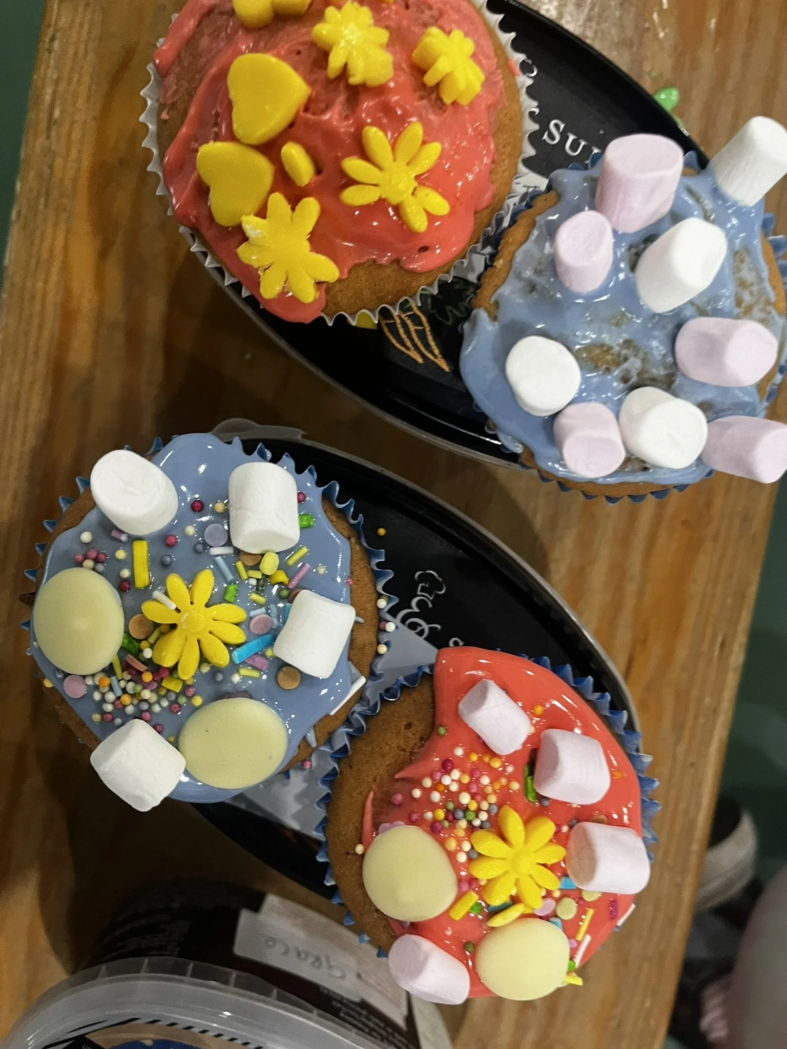 Four decorated cupcakes on a tray, with pink, blue, and red icing, topped with marshmallows, sprinkles, and yellow flower-shaped decorations.