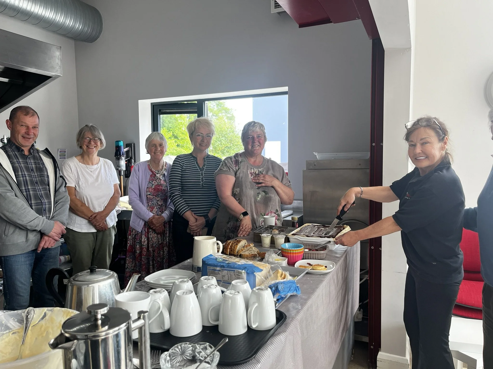 Group of six people standing behind a table with food, some smiling as a woman cuts a cake.